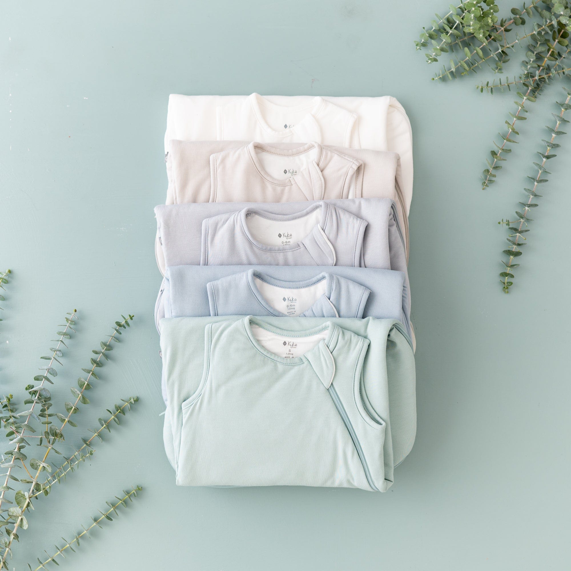 Kyte Baby Sleep Bag TOG 1.0 in core colors stacked staggered one on top of the other on a sage surface