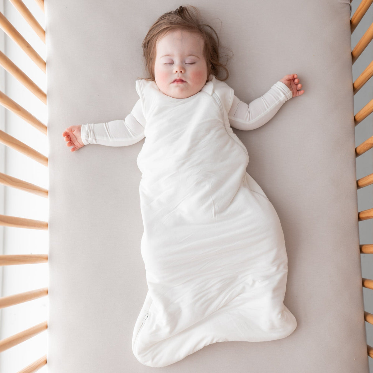 Baby sleeping in crib while wearing Kyte Baby Sleep Bag in Cloud 1.0 with matching Long Sleeve Bodysuit underneath