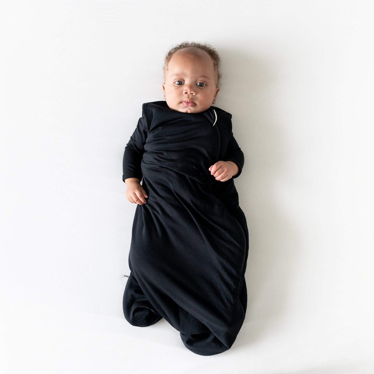 Baby laying on a white surface while wearing Kyte Baby bamboo Sleep Bag in Midnight 2.5 over matching long sleeve bodysuit