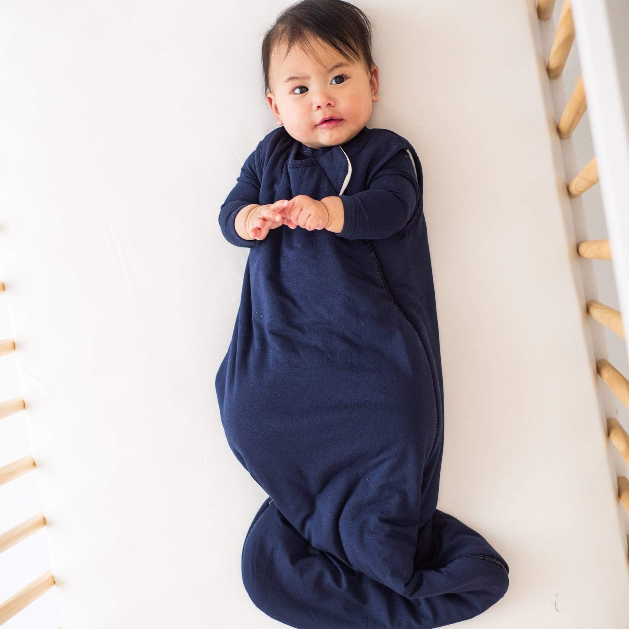 Baby laying in a crib while wearing a Kyte Baby Sleep Bag in Navy 2.5 TOG and a matching long sleeve bodysuit underneath