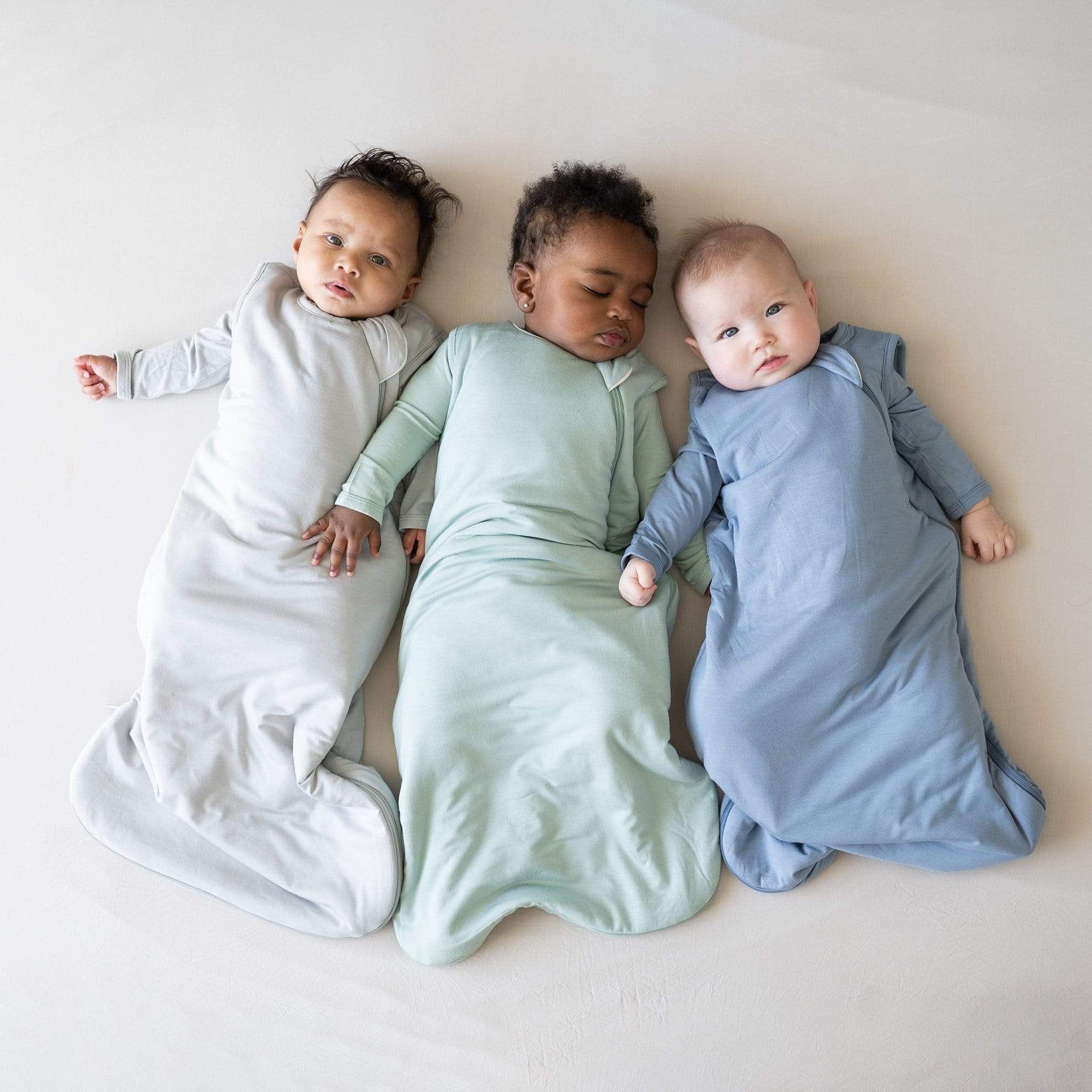 Three babies laying next to each other wearing Kyte Baby sleep bags TOG 2.5 in Storm, Sage and Slate