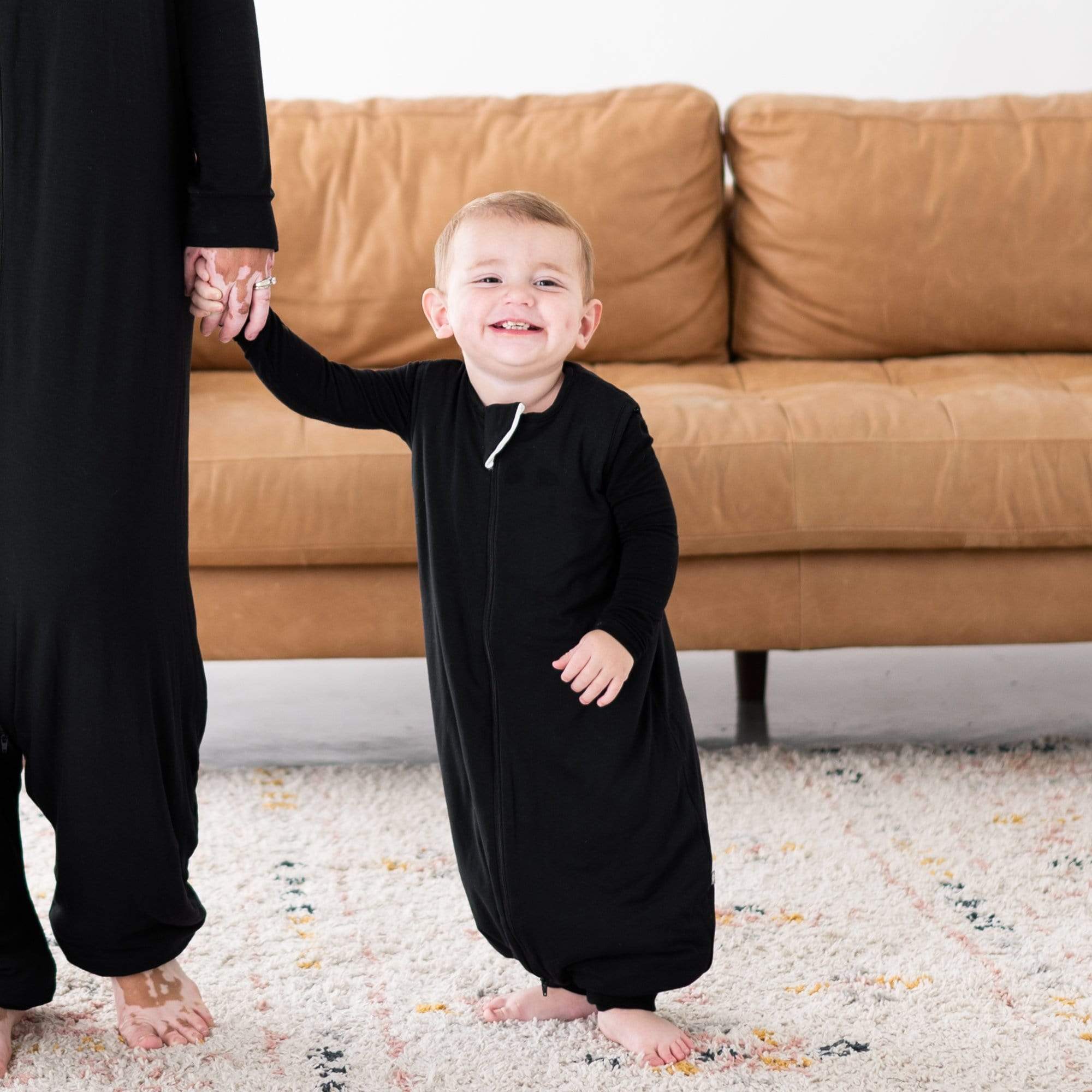 Smiling toddler in a Sleep Bag Walker in Midnight 1.0 holding hands with an adult in a matching Adult Sleep Bag Walker. They are standing on a light-colored rug in front of a tan couch