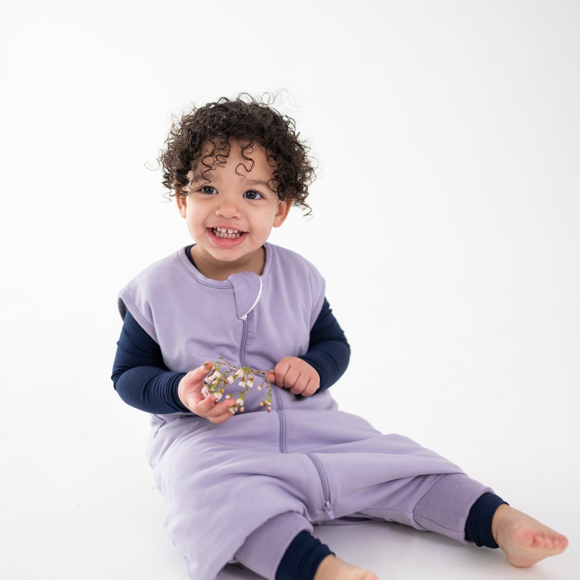Child sitting on the floor holding flowers wearing Kyte Baby bamboo Sleep Bag Walker in Taro 1.0