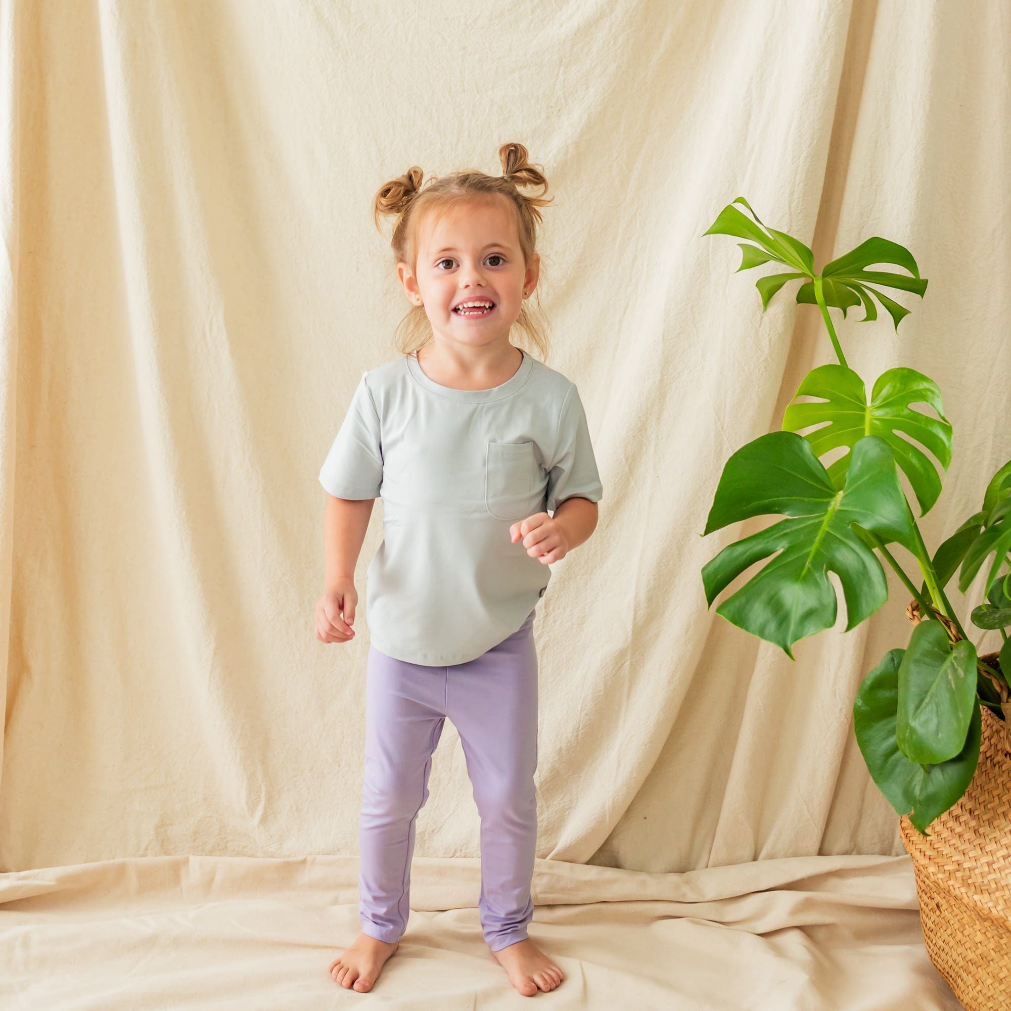 Child standing next to a plant while wearing Toddler Leggings in Taro and a tee in Sage
