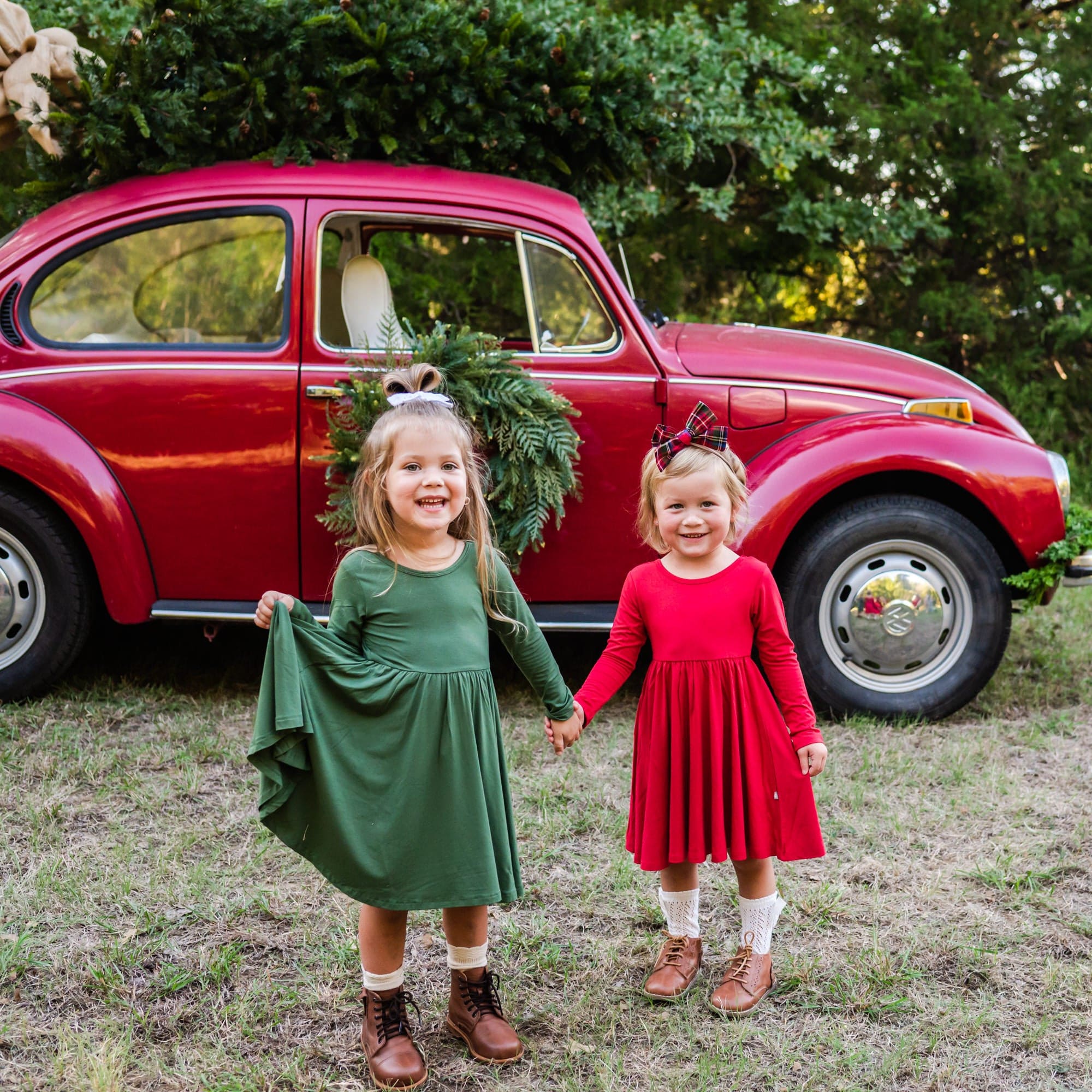 Two girls standing together holding hands outside wearing green and red Long Sleeve Twirl Dresses standing in front of a red car