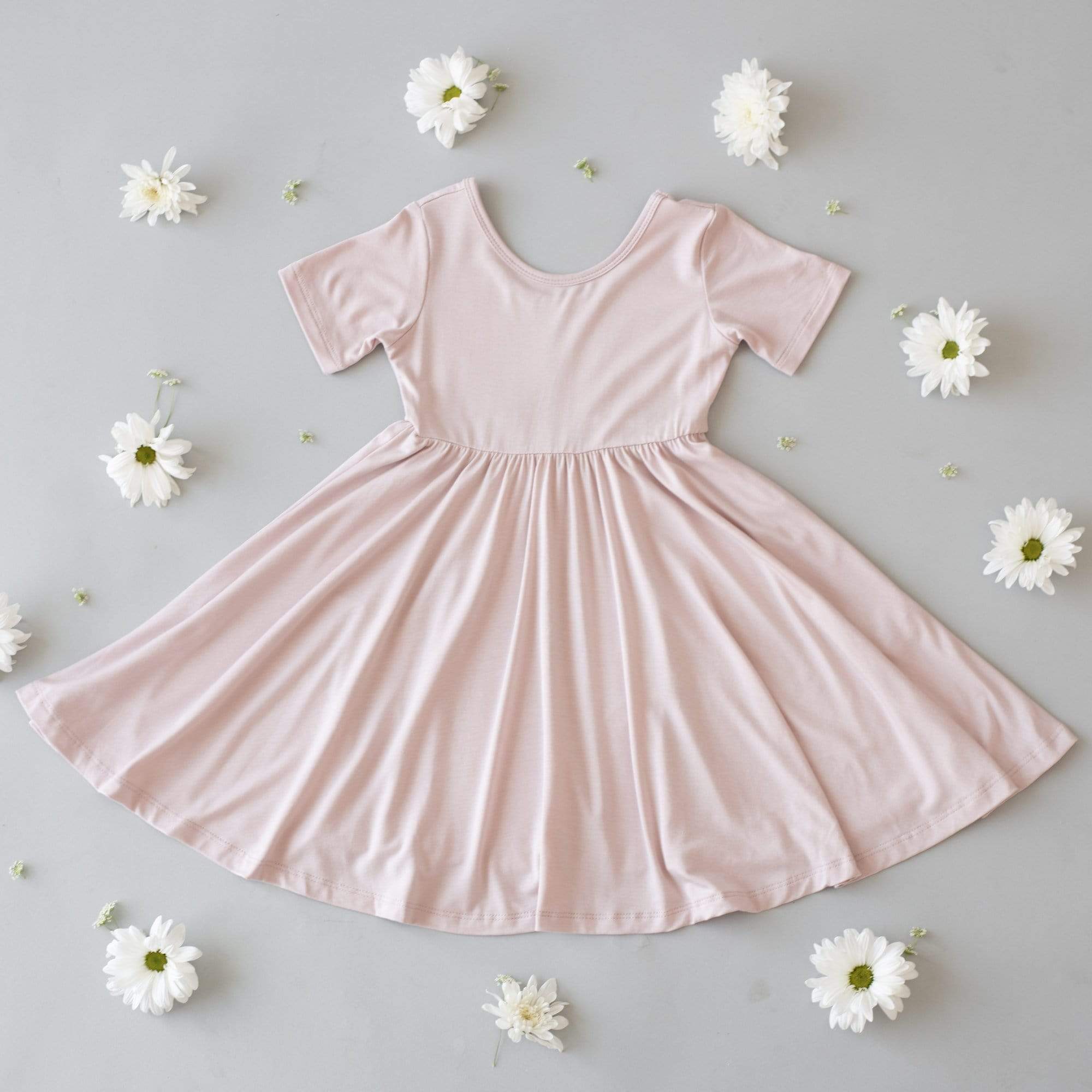 Flat lay of a Twirl Dress in Blush on a gray surface with white flowers spread around it