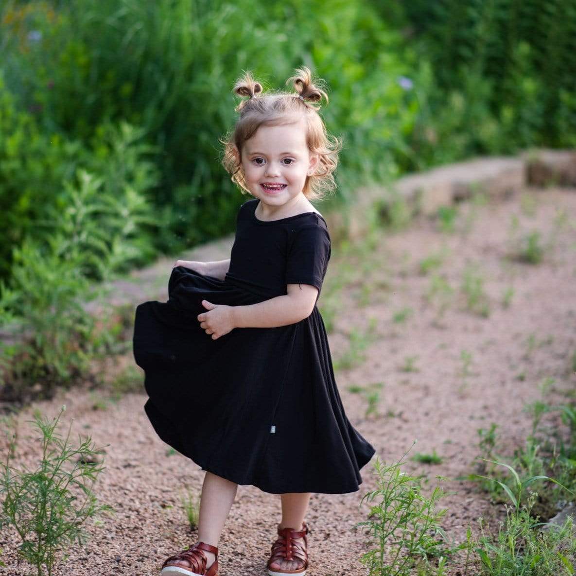 Toddler wearing Kyte Baby Twirl Dress in Midnight on a wooded walkway