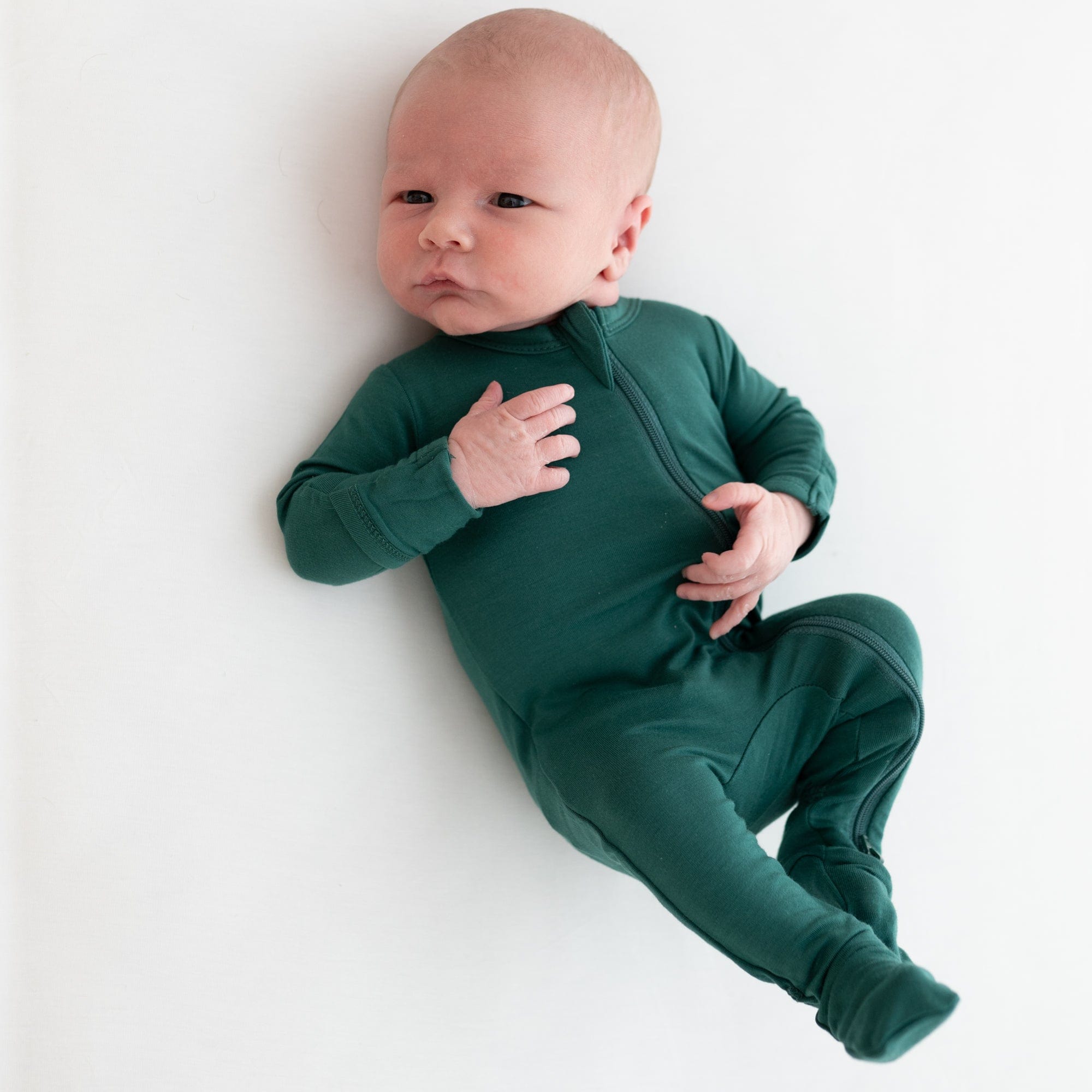Newborn laying on a white surface while wearing a Zippered Footie in Emerald