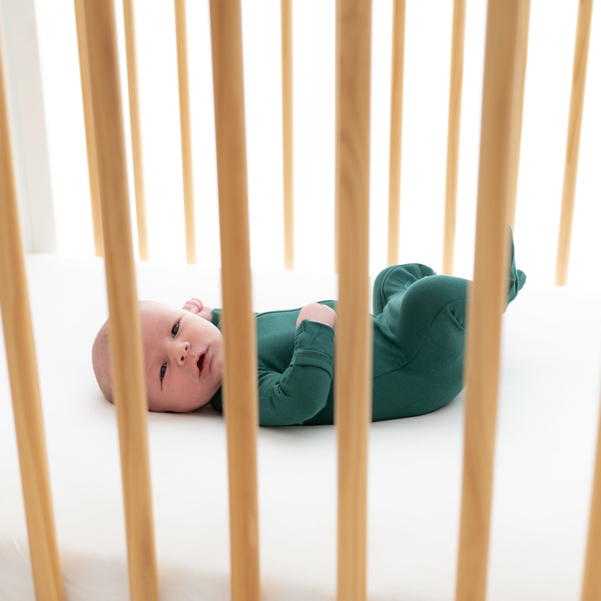 Newborn laying in a wooden crib wearing Kyte Baby Zippered Footie in Emerald
