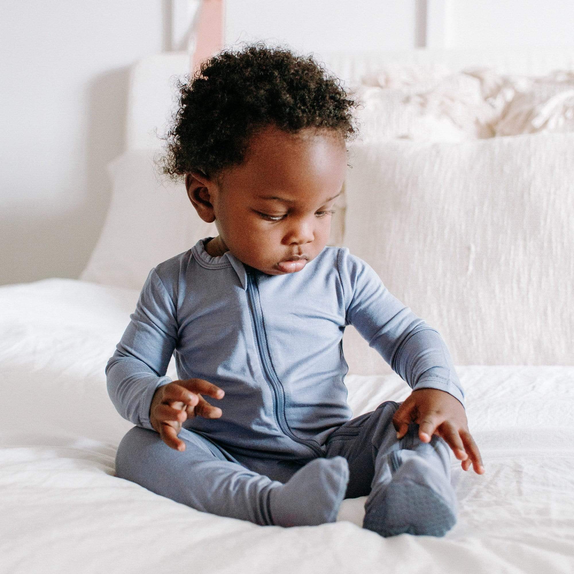 Baby sitting on a white bed and wearing a Zippered Footie in Sage