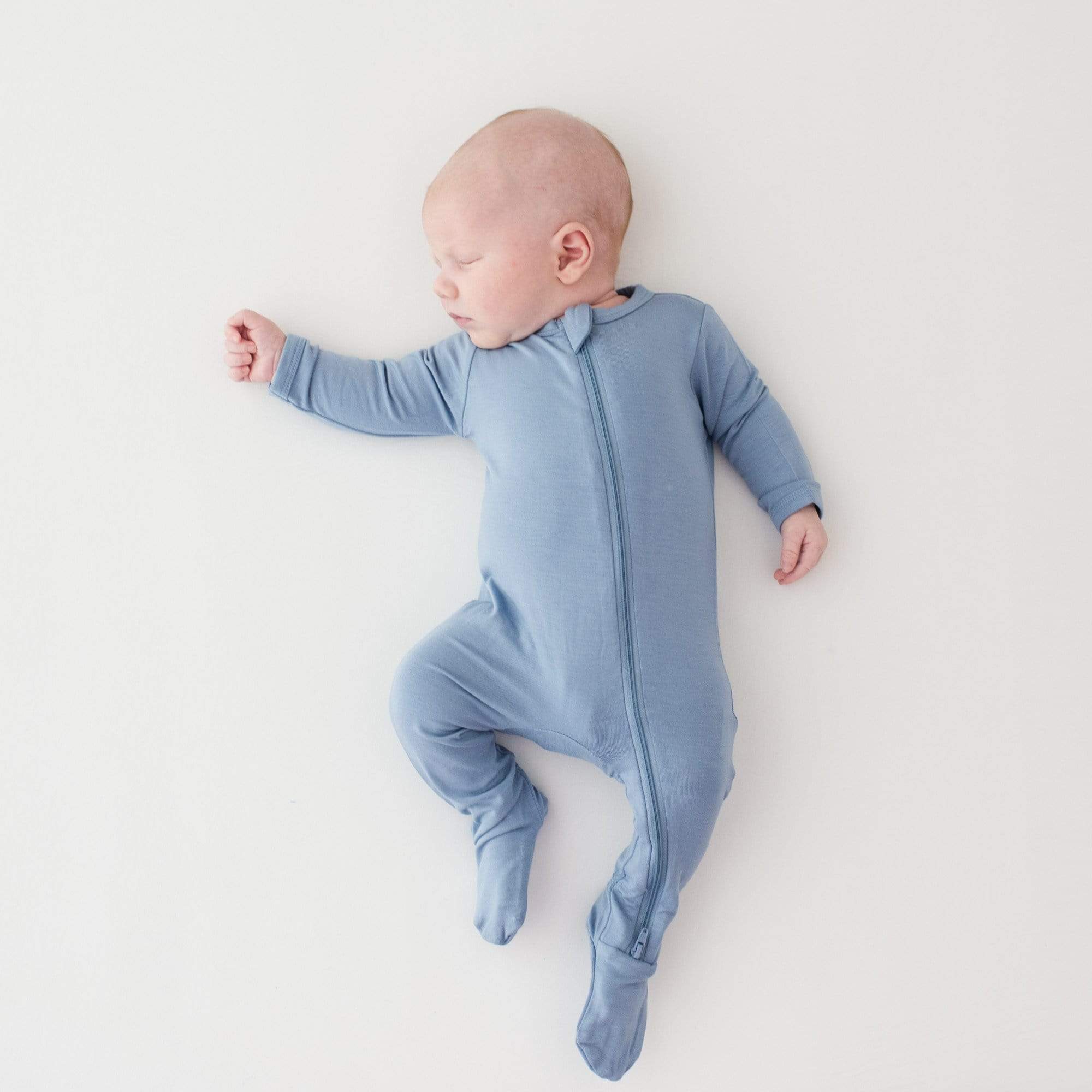 Sleeping infant laying on a white surface while wearing a Kyte Baby Zippered Footie in Slate
