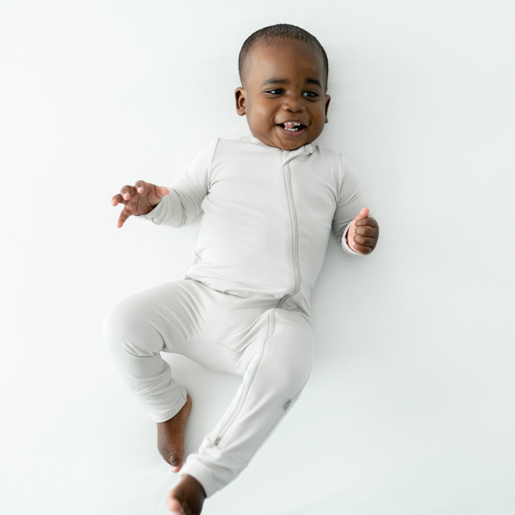 Baby laying on a white surface while wearing Kyte Baby Zippered Romper in Oat