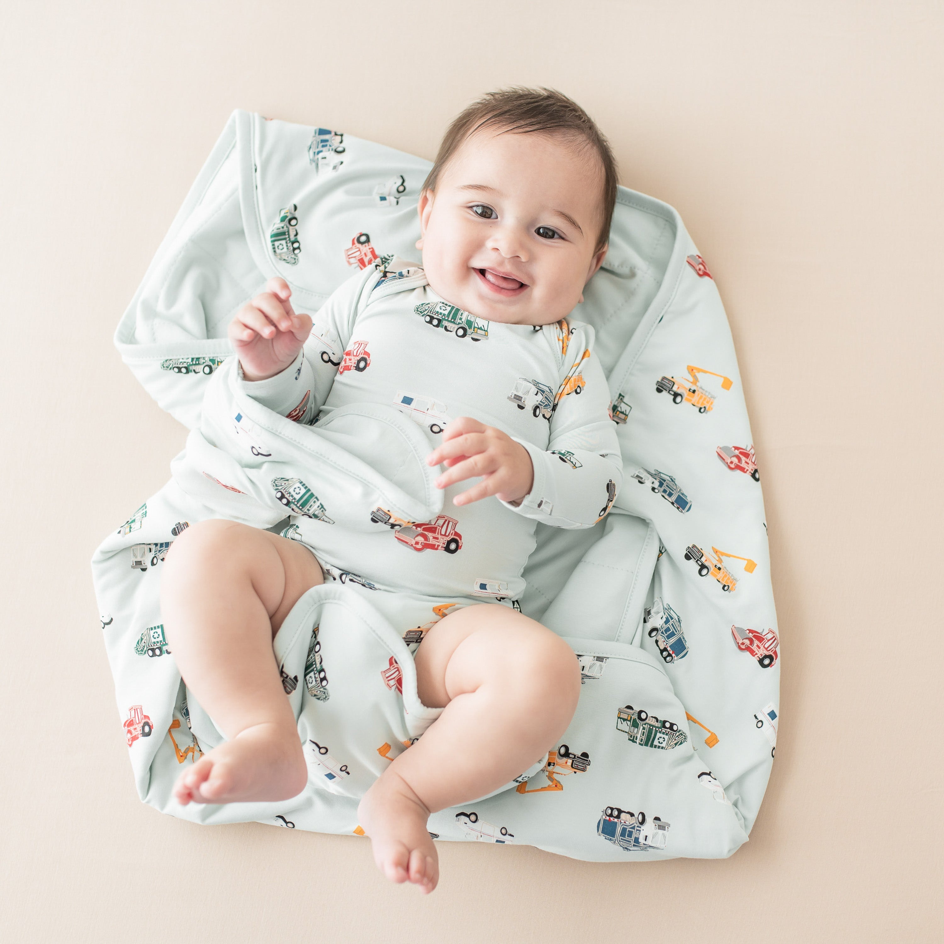 Baby wearing a long sleeve bodysuit with a vehicle pattern laying on a matching printed blanket