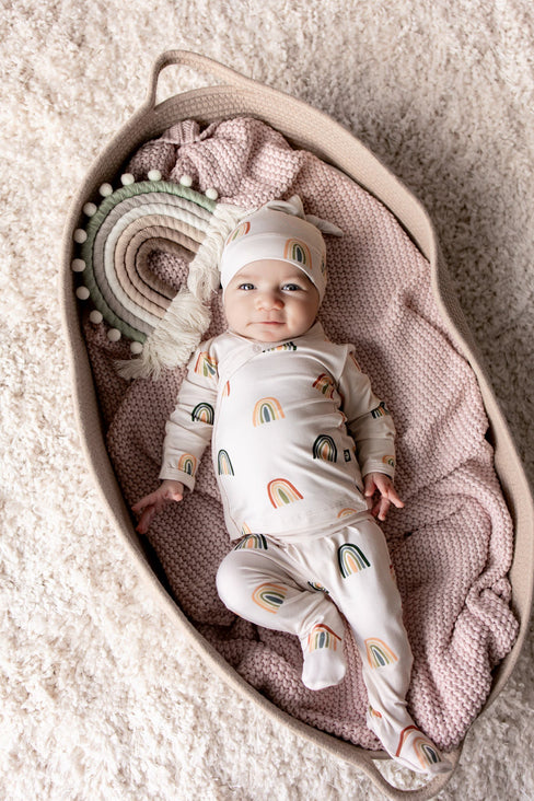 Baby lying in a basket over a pink blanket wearing Take Me Home Set with Cap in Retro Rainbow print with a retro rainbow prop