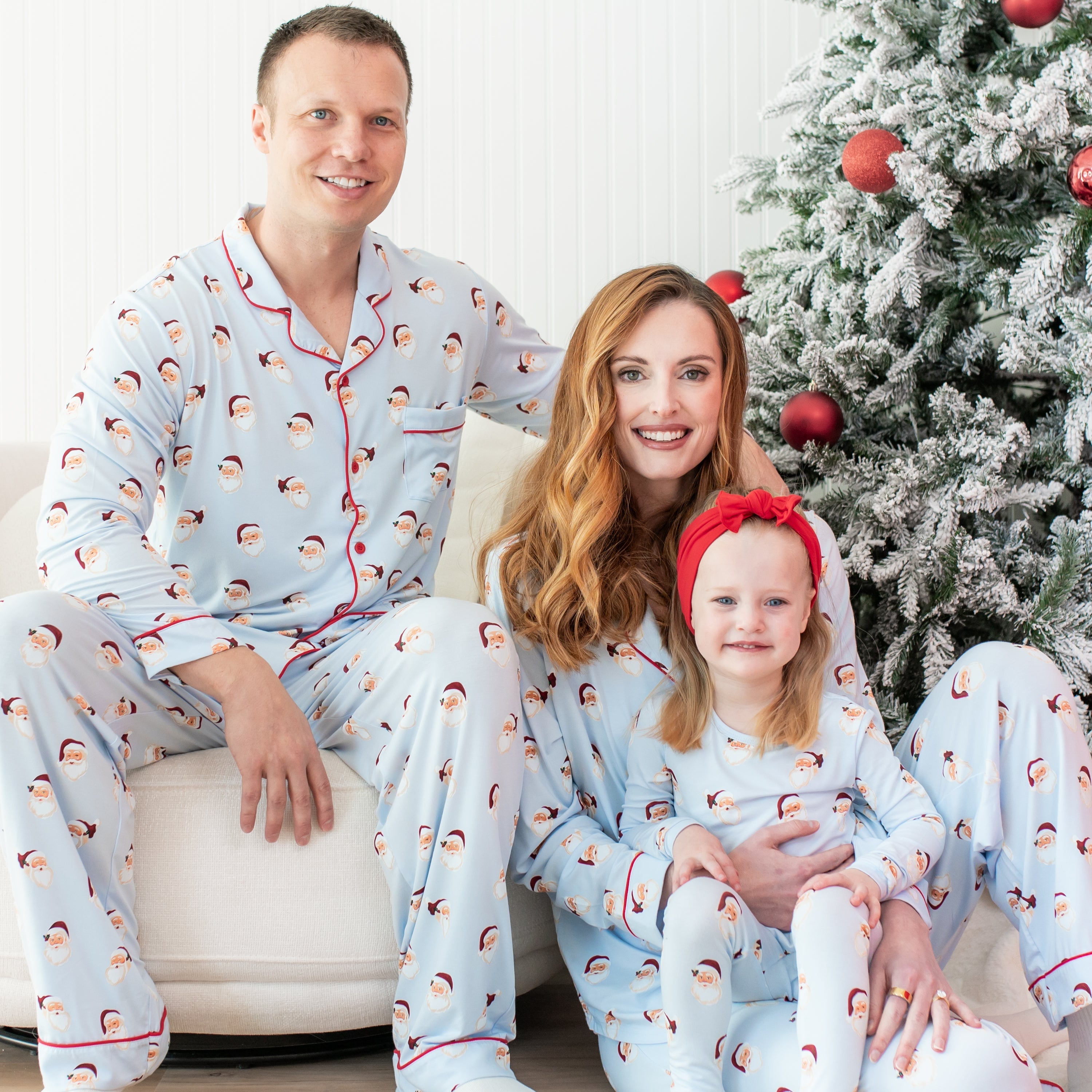 Family of 3 sitting near a decorated christmas tree wearing matching family holiday pajamas with a vintage santa print