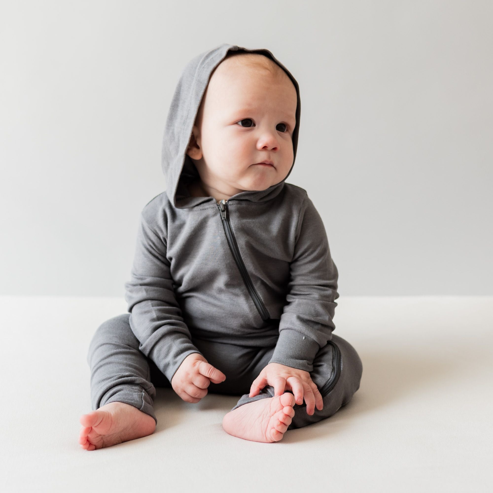 Baby sitting wearing bamboo jersey hooded zipper romper in the color charcoal 