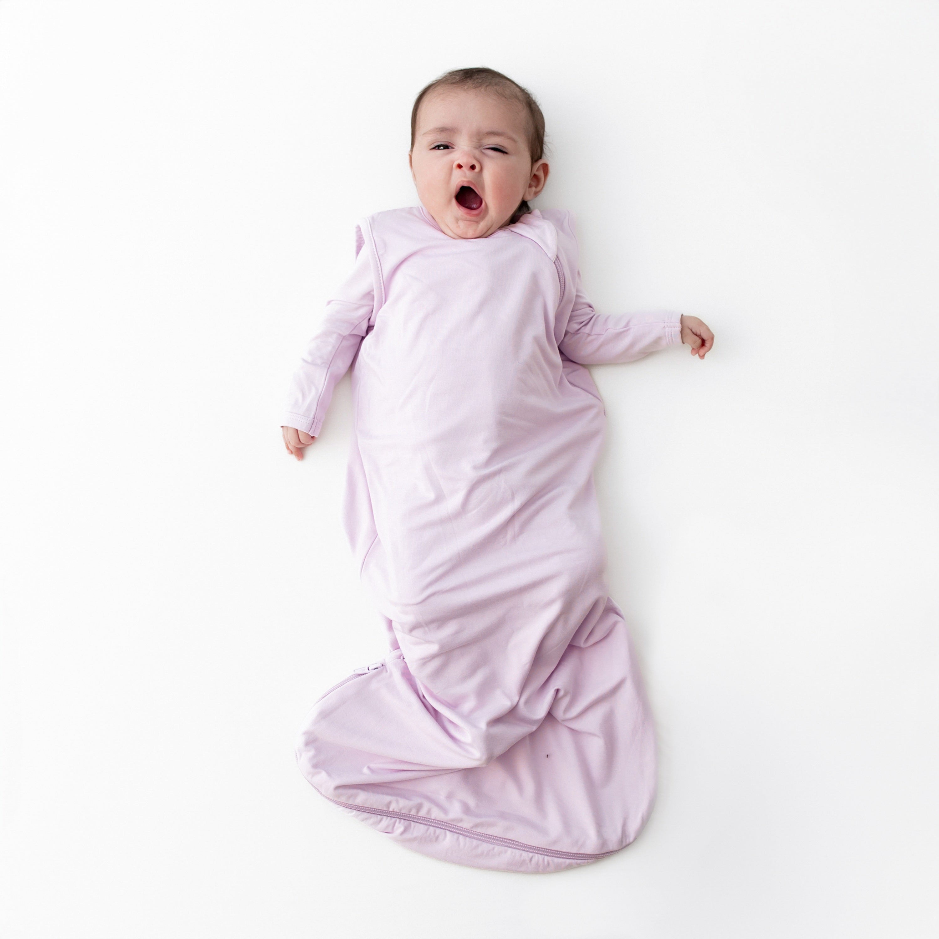 Yawning baby laying on a white surface wearing the Sleep Bag in Thistle 0.5 with matching romper