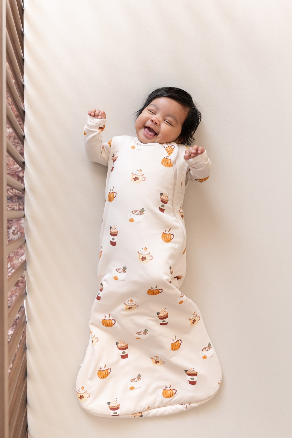 Baby smiling in Sleep Bag with Pumpkin Spice Latte 1.0 print on a beige crib sheet