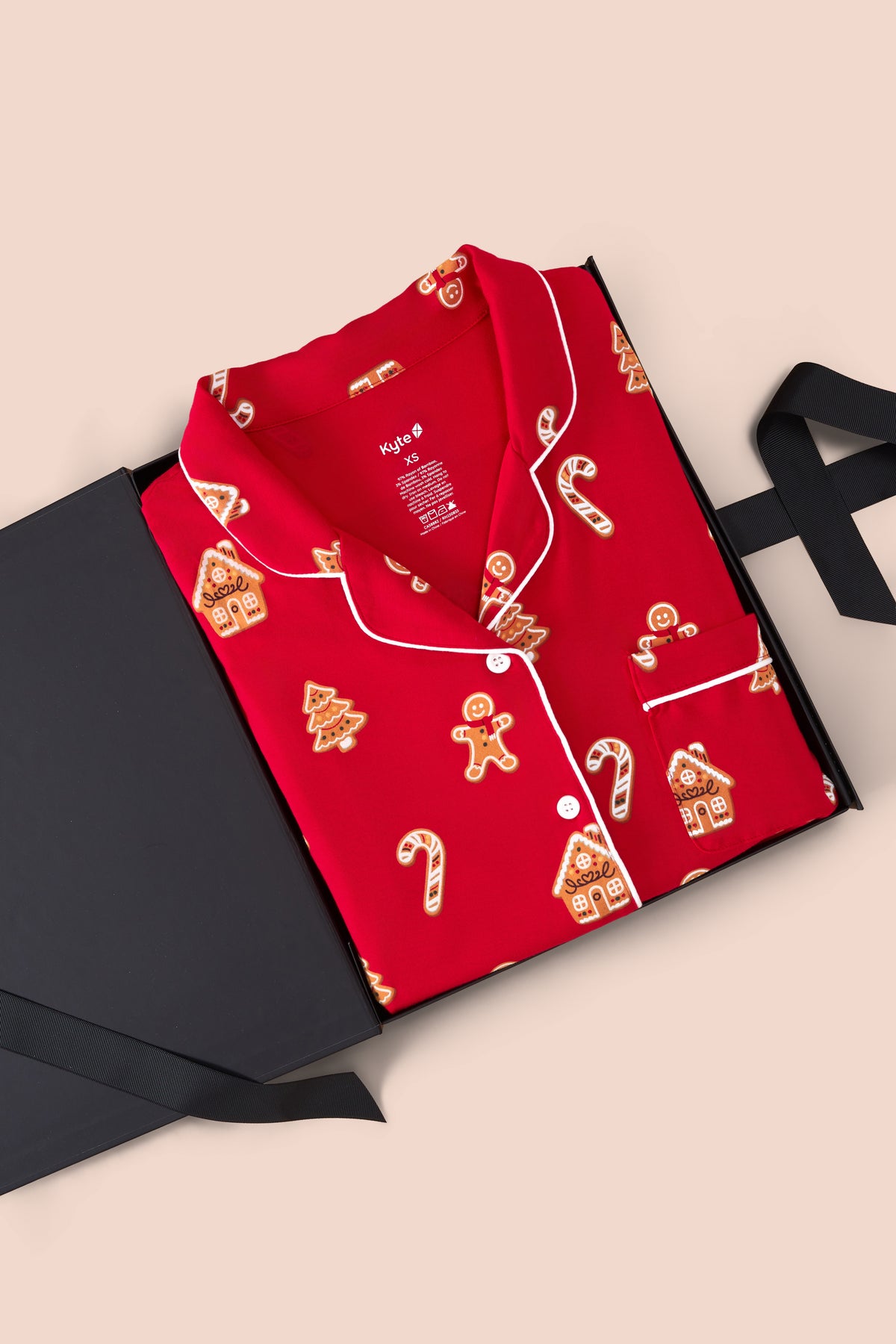A red long-sleeve pajama shirt with white piping and a holiday print of gingerbread houses, trees, and candy canes, presented in an open black gift box on a beige background.