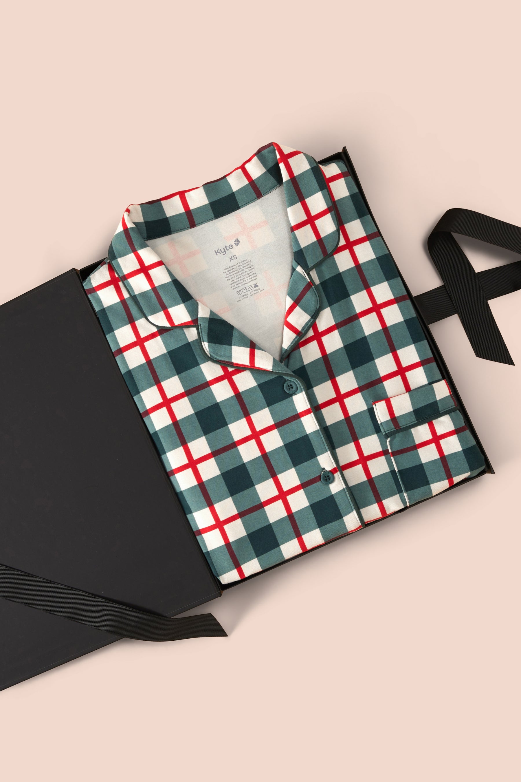 A folded plaid pajama shirt in green, white, and red is presented in a black gift box with a black ribbon on a light beige background.