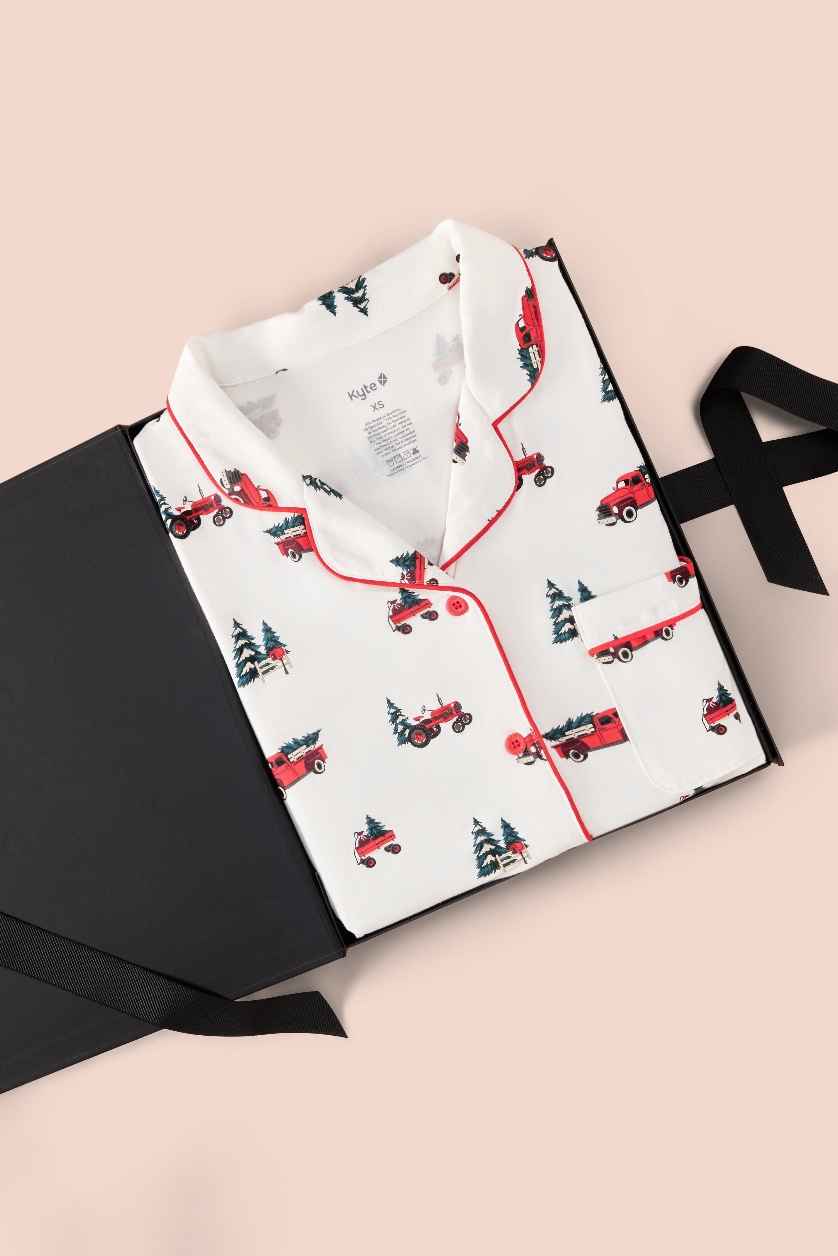 A folded white pajama shirt with red piping and a repeating print of red pickup trucks and dark green Christmas trees, displayed on a black gift box with a black ribbon against a pale pink background.