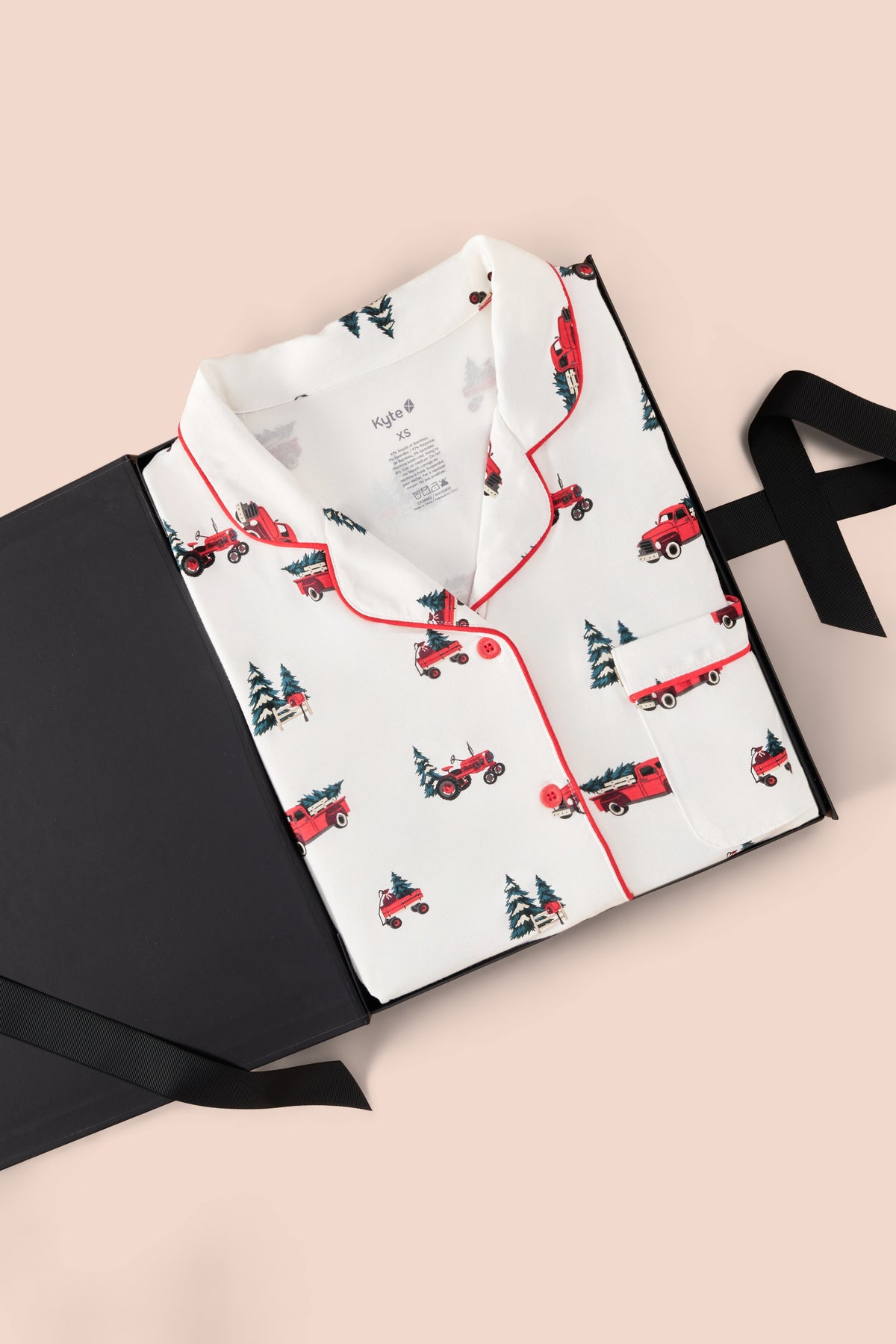 A folded white pajama shirt with red piping and a repeating print of red pickup trucks and dark green Christmas trees, displayed on a black gift box with a black ribbon against a pale pink background.