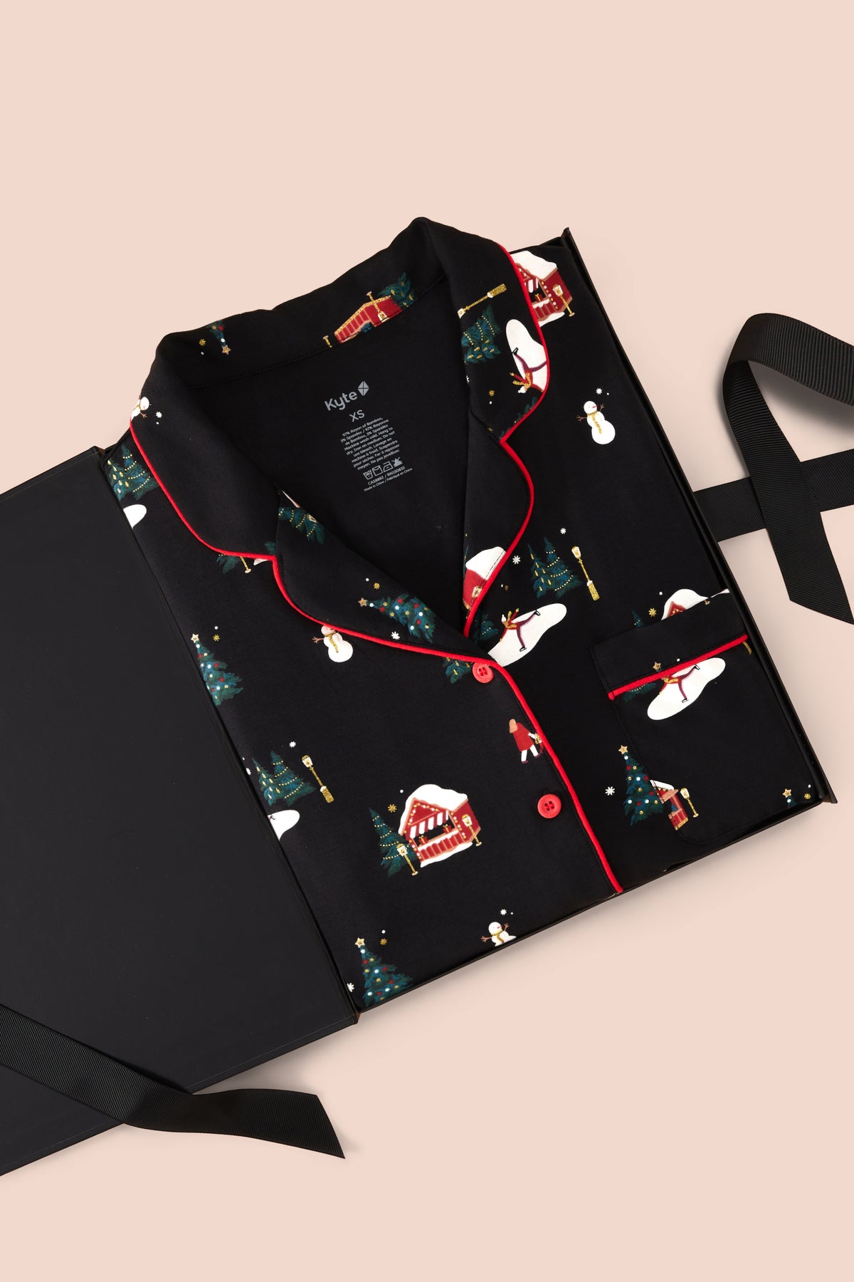 A black pajama set with a Christmas-themed print, featuring red piping and red buttons, presented on a black gift box with a ribbon against a light beige background.