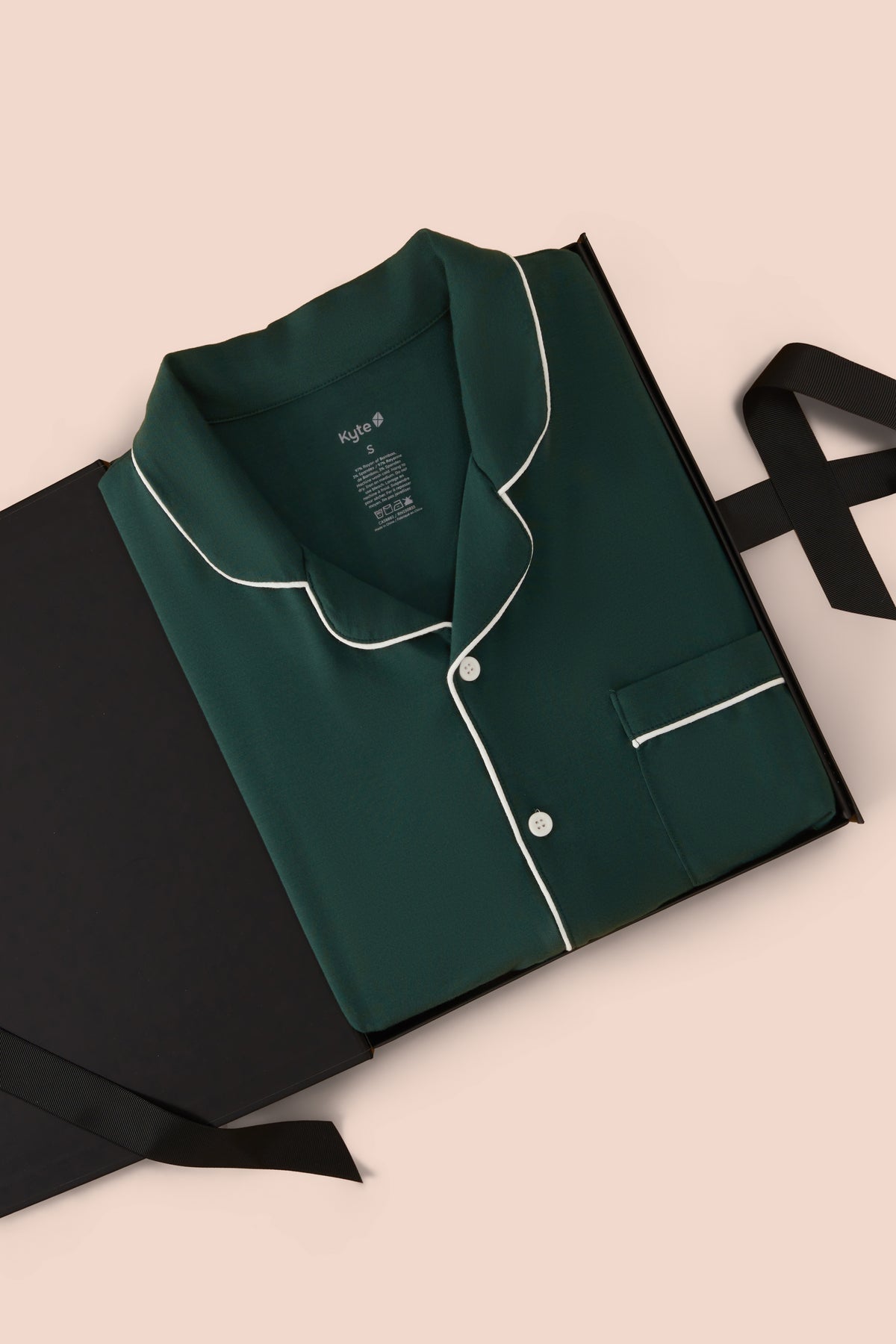 A dark green pajama shirt with white piping trim, folded and presented in a black gift box with a black ribbon on a light beige background.