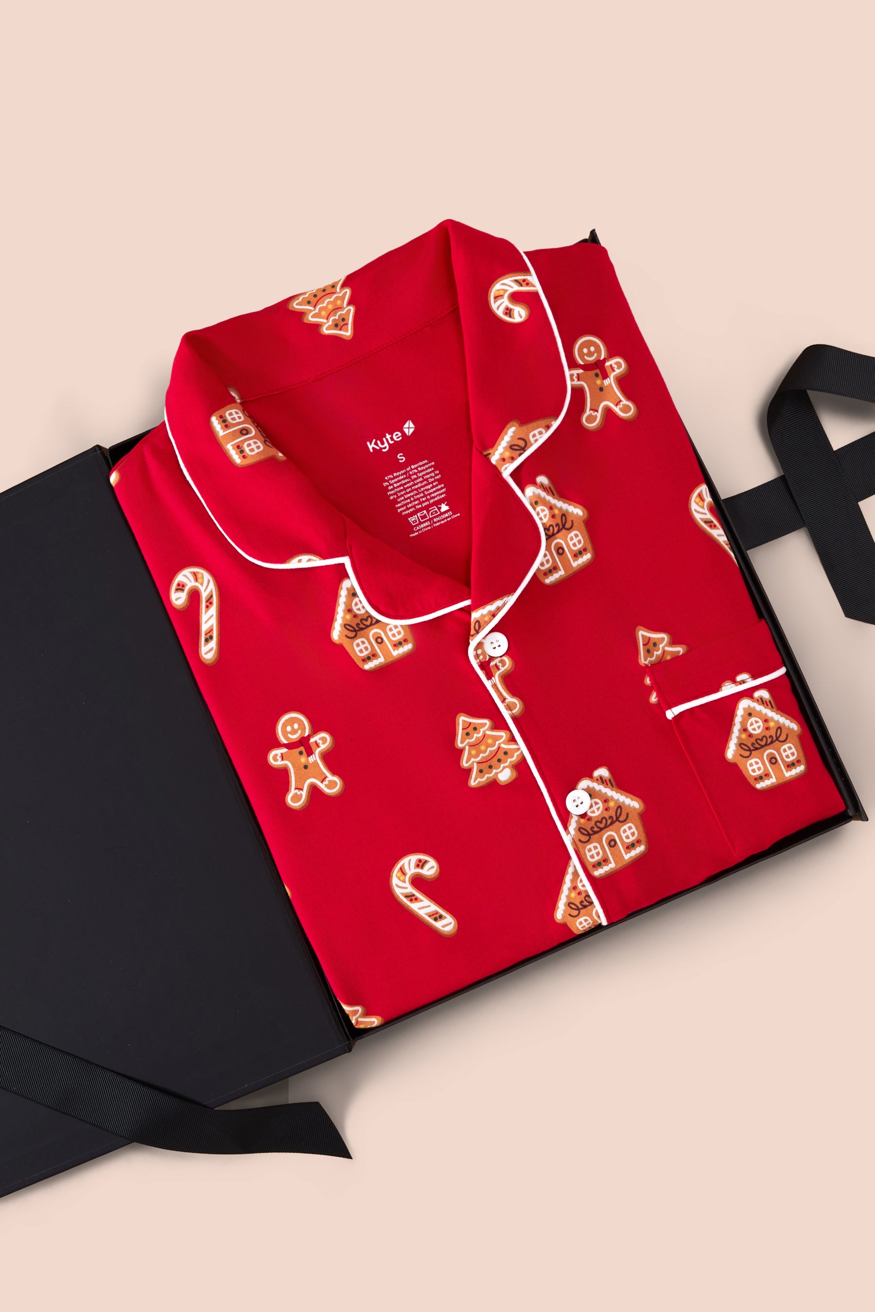 A red long-sleeve pajama shirt with white piping and gingerbread-themed prints, presented in an open black gift box on a light beige background.