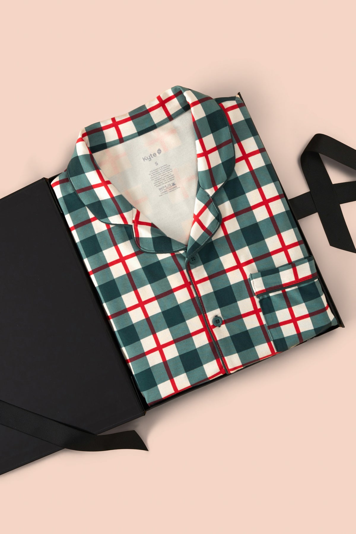 A folded long-sleeve plaid pajama shirt in green, white, and red, presented in a black gift box with a black ribbon on a solid pale pink background.
