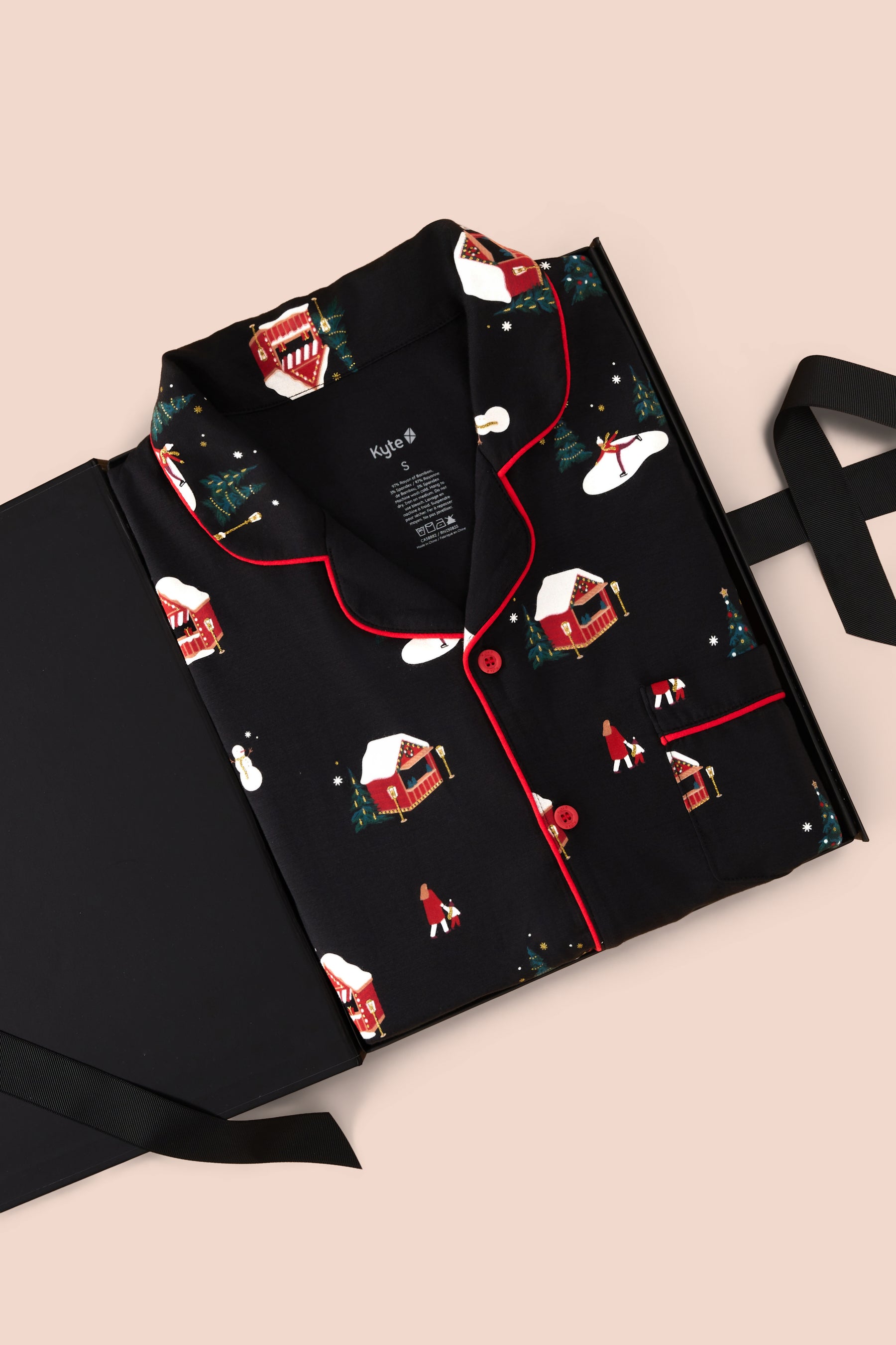 A folded black pajama set with a Christmas village print, featuring red piping and a button-down front, displayed on a black gift box with a ribbon against a light beige background.