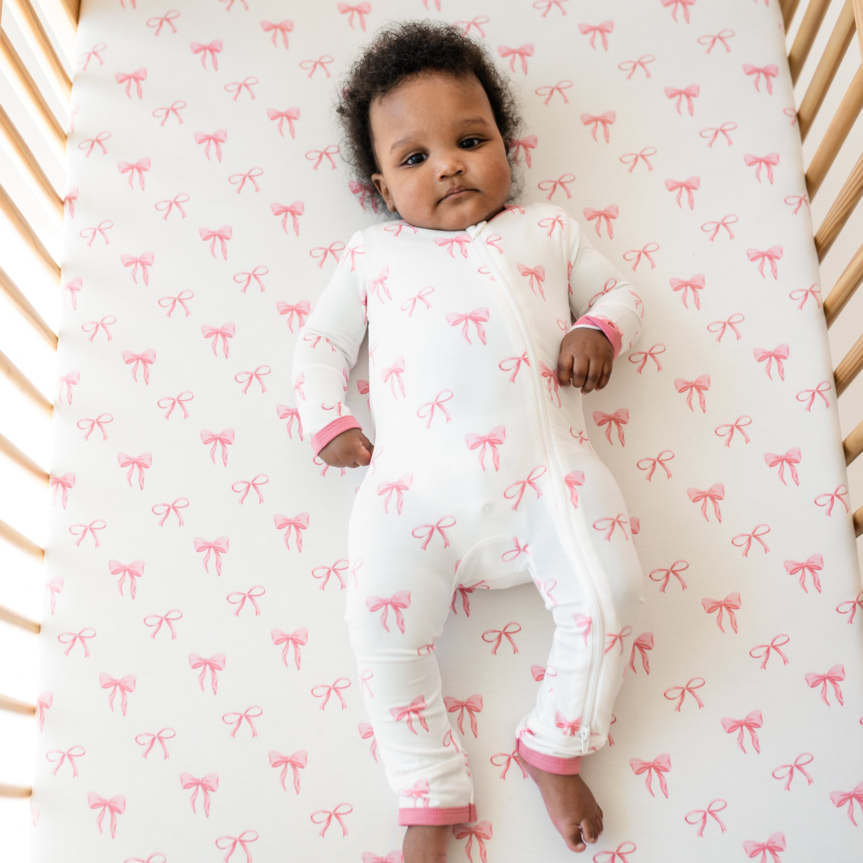Baby in Zippered Romper pajamas laying on Crib Sheet in Bow pattern