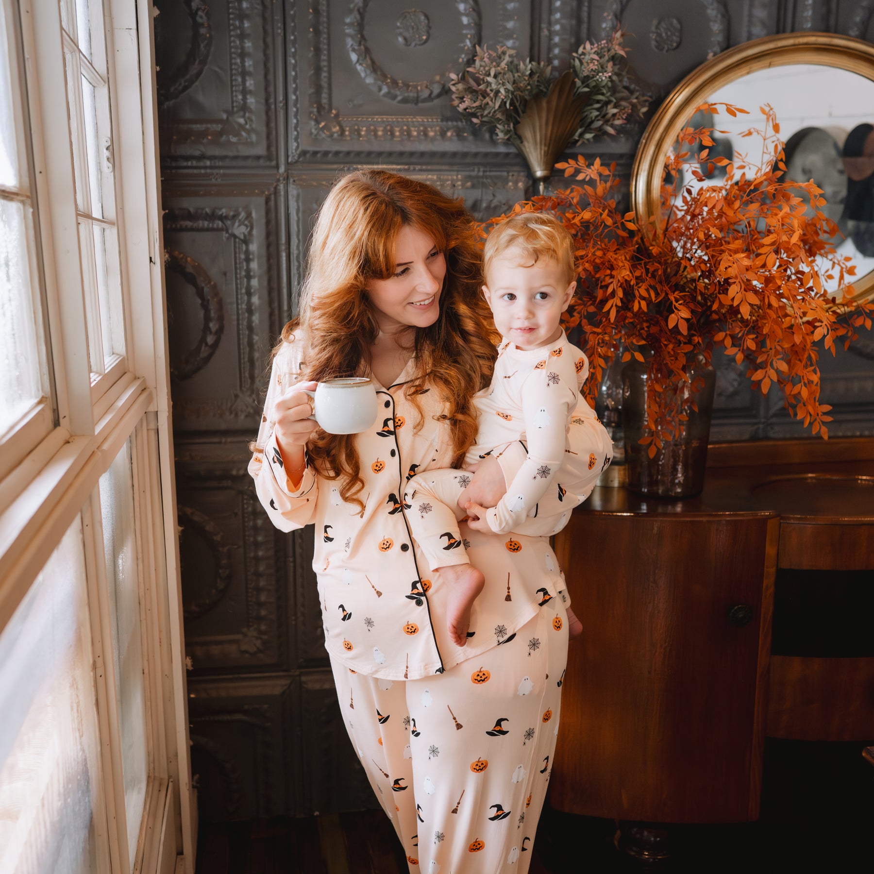 Mother wearing the Long-Sleeved Women's Pajama Set in Bewitched holding her son who is in a matching zippered romper and holding a cup of coffee in the other hand