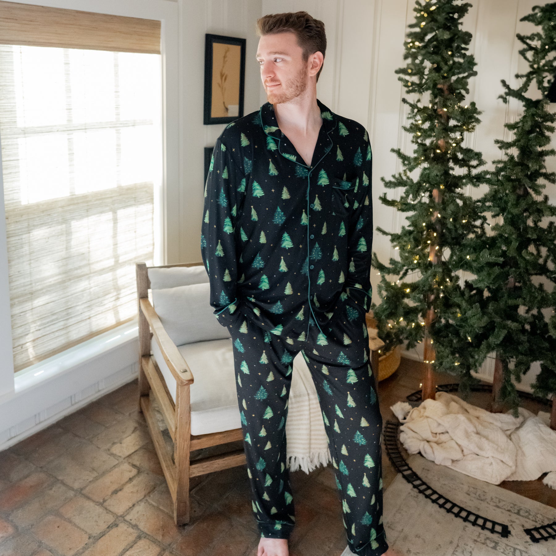Male model standing with hands in the pockets of the Men's Long Sleeve Pajama Set in Twinkle Tree in front of a wooden chair and Christmas tree