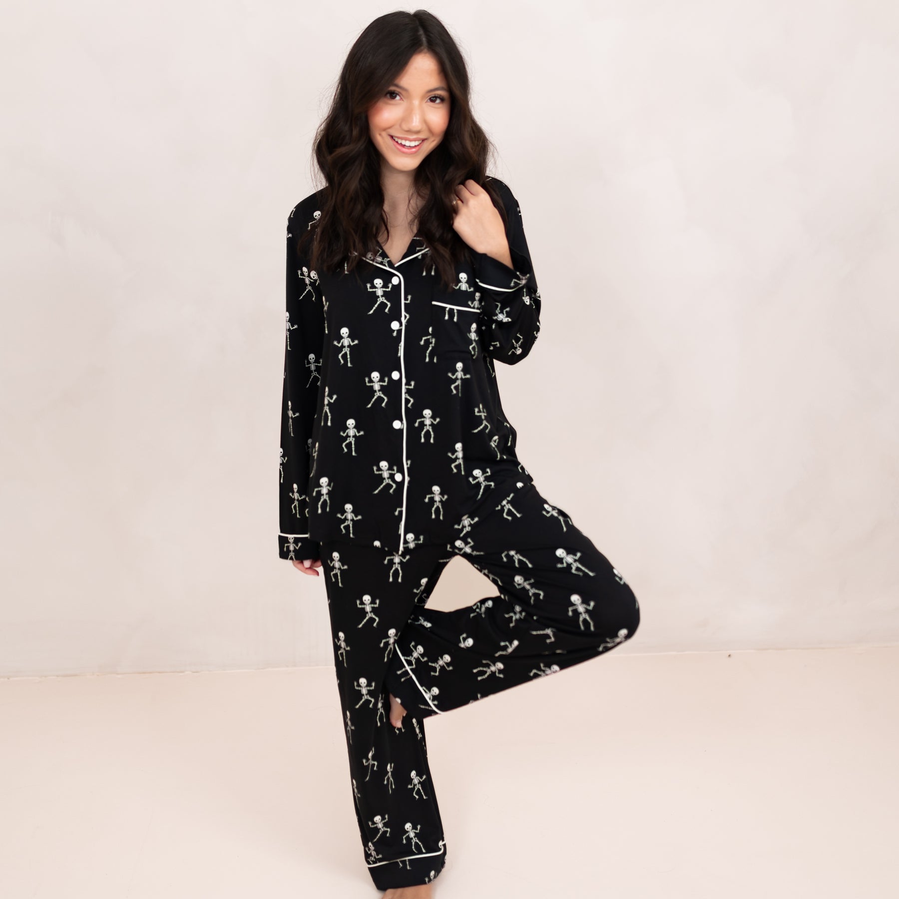 Smiling model standing on one leg with the other leg bent and resting on her knee wearing the Long-Sleeved Women's Pajama Set in Skeleton in front of a light beige backdrop