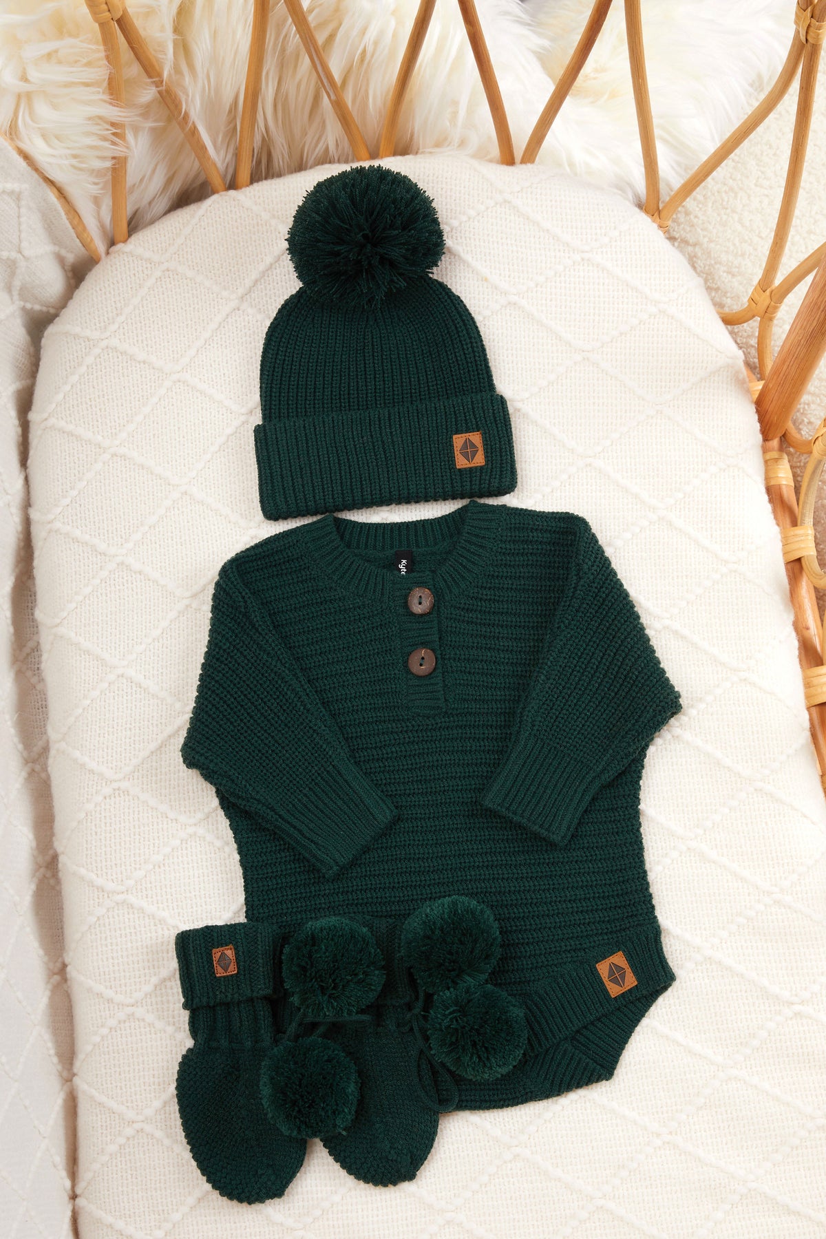 Chunky knit pom beanie, chunky knit romper and knit booties in cypress laid in a crib