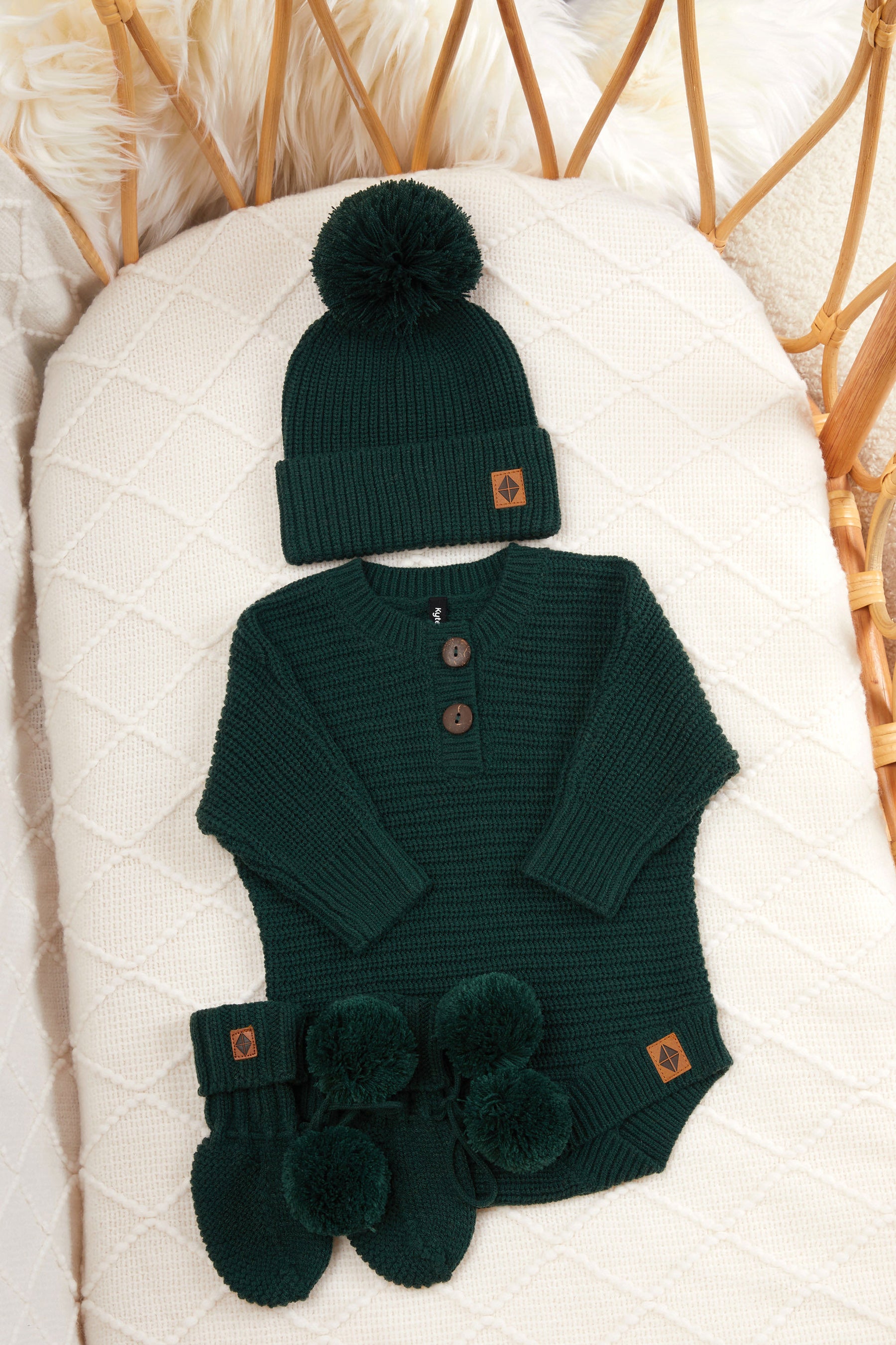 Chunky knit pom beanie, chunky knit romper and knit booties in cypress laid in a crib