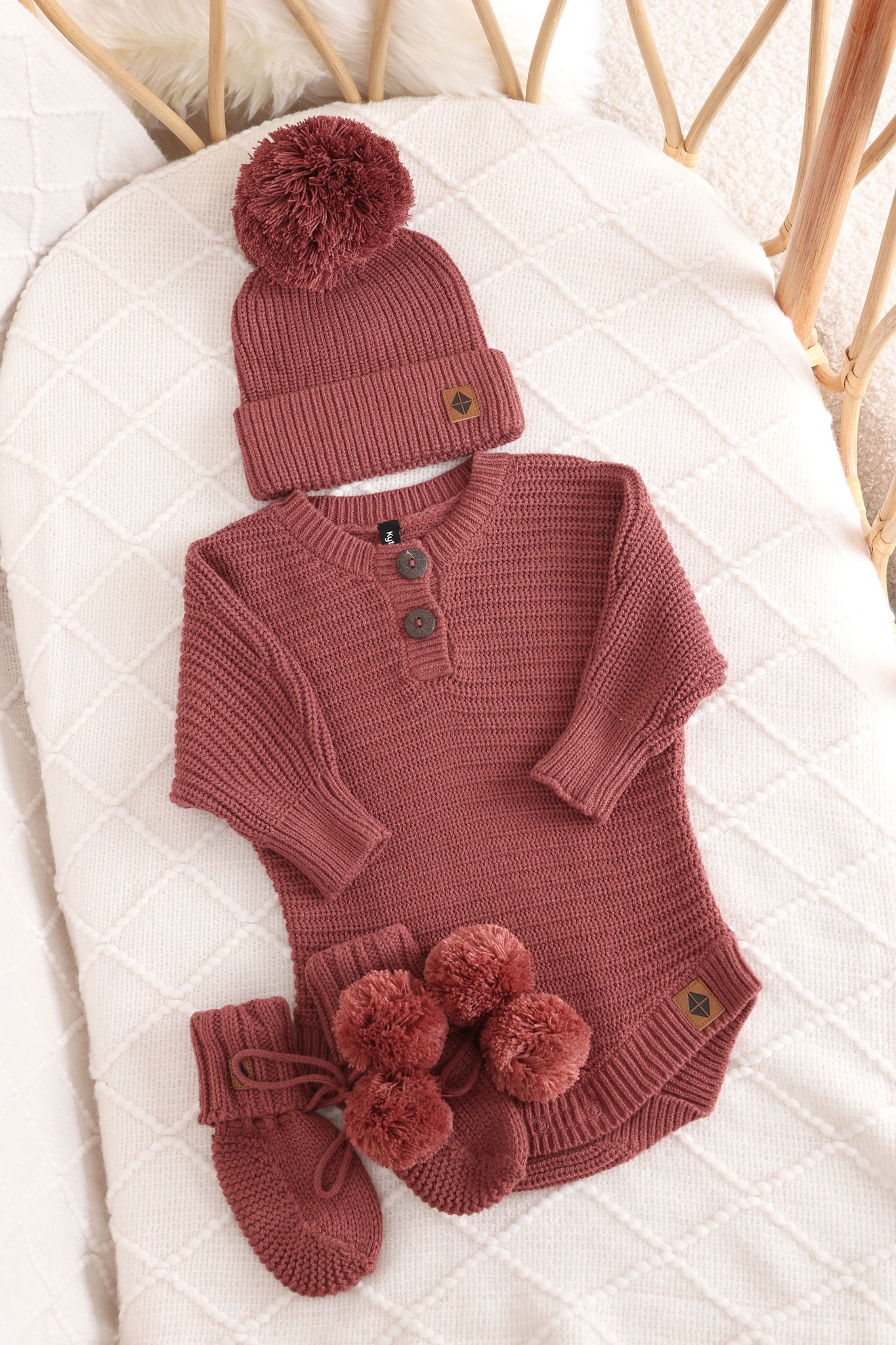 Chunky knit pom beanie, chunky knit romper and knit booties in ember laid in a crib
