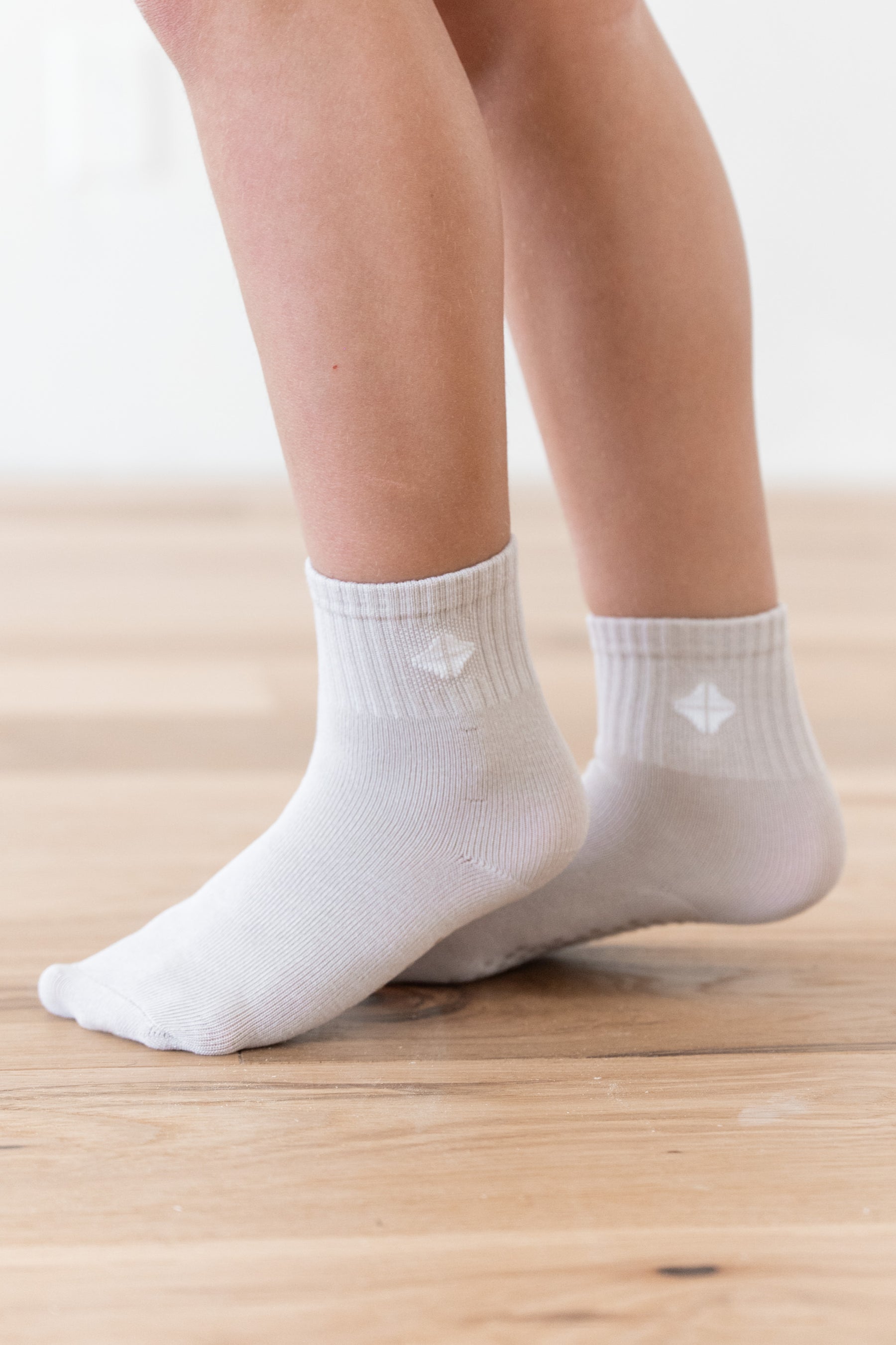 White ankle socks worn on a child's feet, standing on a light wooden floor against a bright white background.