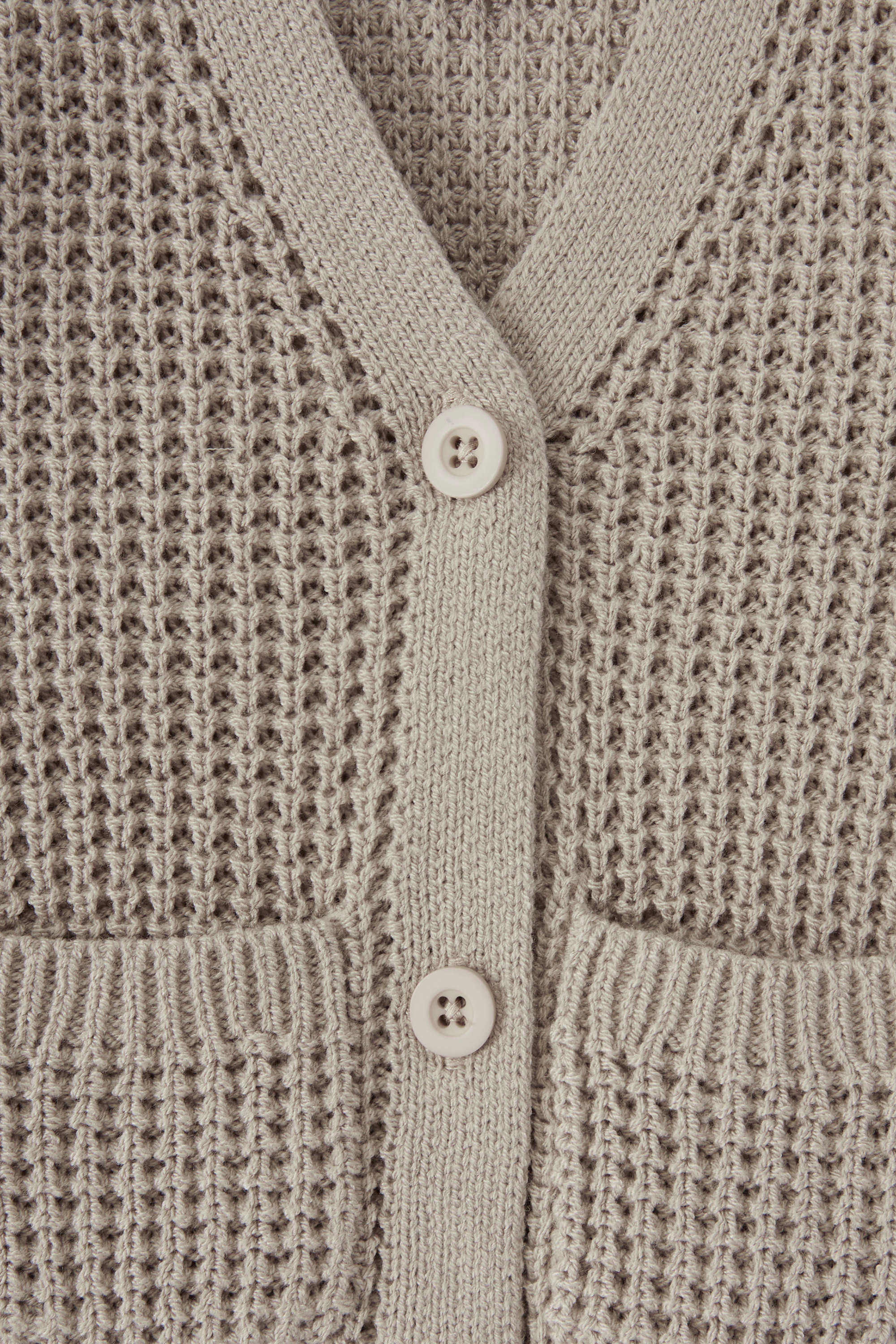 Close-up of a textured knit cardigan in a warm taupe-beige color, featuring a waffle-weave pattern, ribbed trim, and two white buttons.