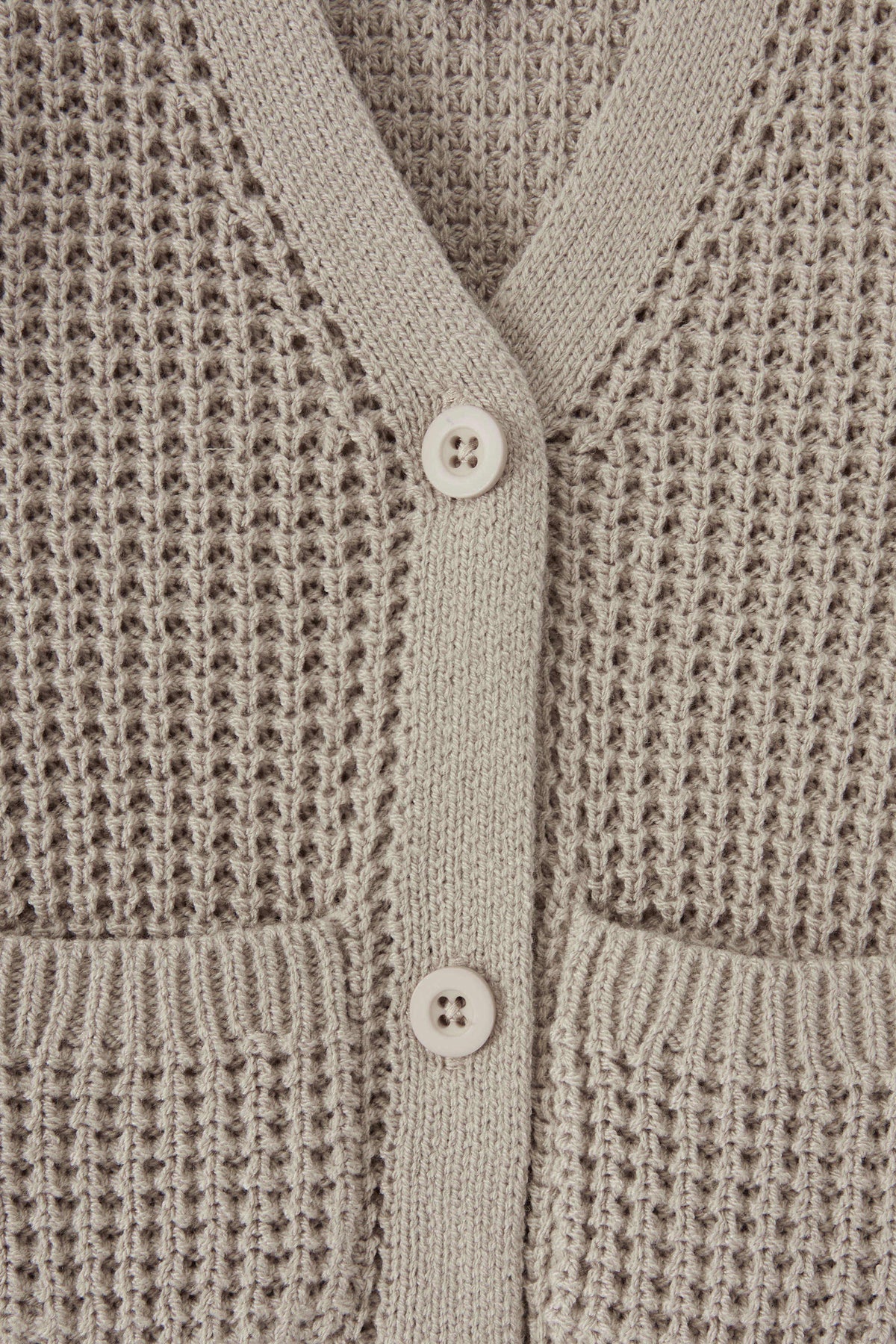 Close-up of a textured knit cardigan in a warm taupe-beige color, featuring a waffle-weave pattern, ribbed trim, and two white buttons.