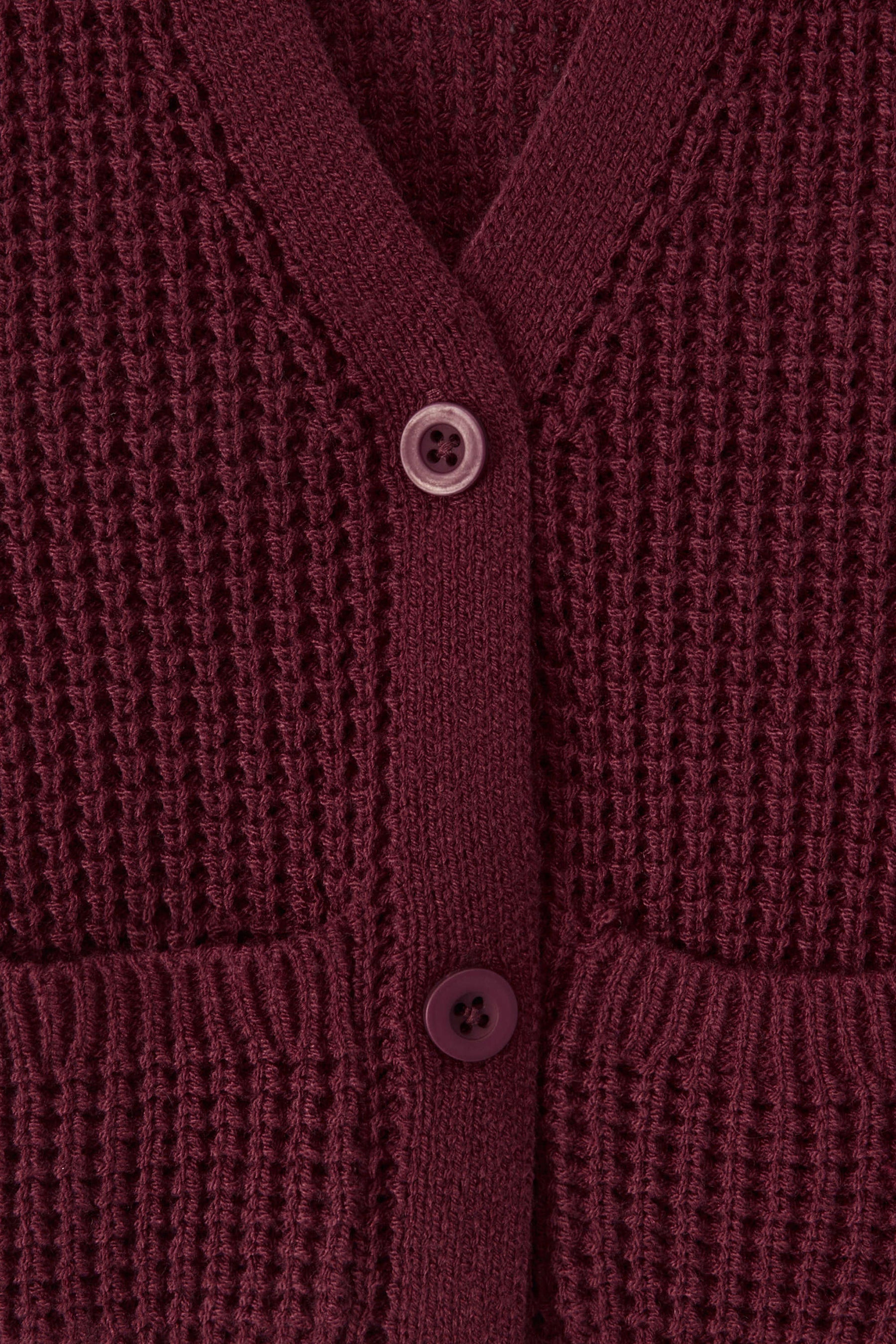 Close-up of a textured knit cardigan in a burgundy color, featuring a waffle-weave pattern, ribbed trim, and two white buttons.