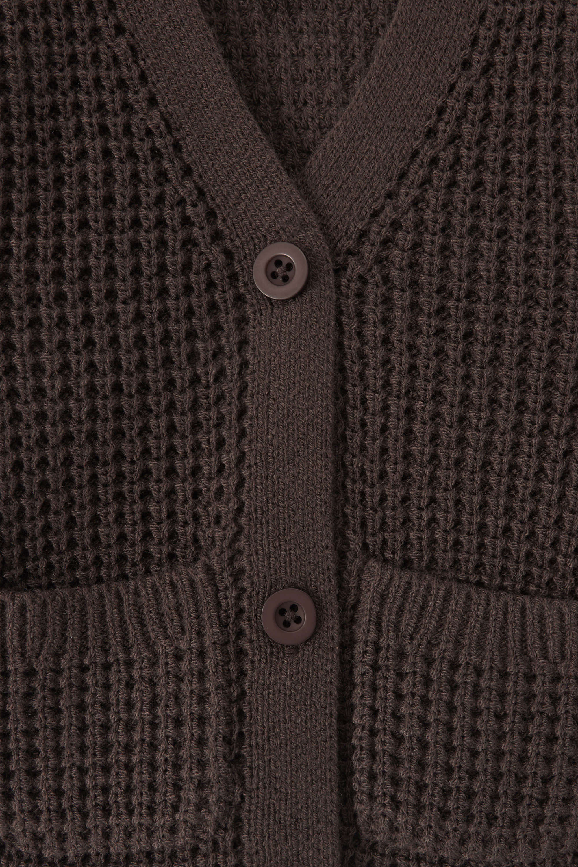 Close-up of a textured knit cardigan in a dark brown color, featuring a waffle-weave pattern, ribbed trim, and two white buttons.