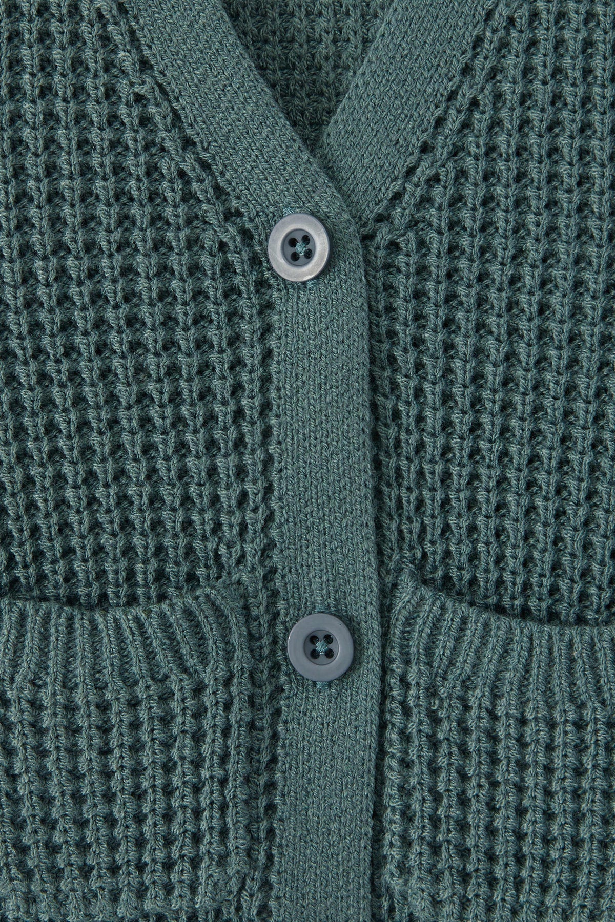 Close-up of a textured knit cardigan in a green color, featuring a waffle-weave pattern, ribbed trim, and two white buttons.
