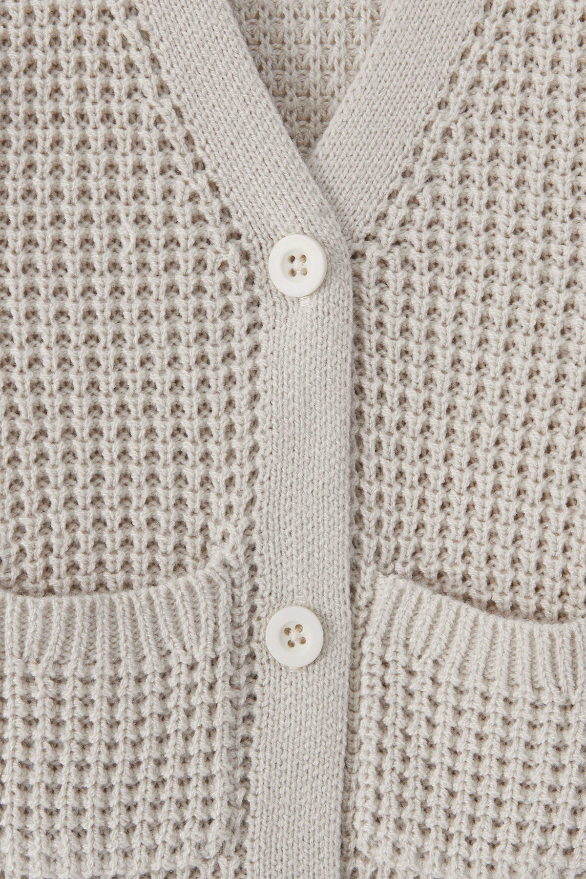 Close-up of a textured knit cardigan in an oat color, featuring a waffle-weave pattern, ribbed trim, and two white buttons.