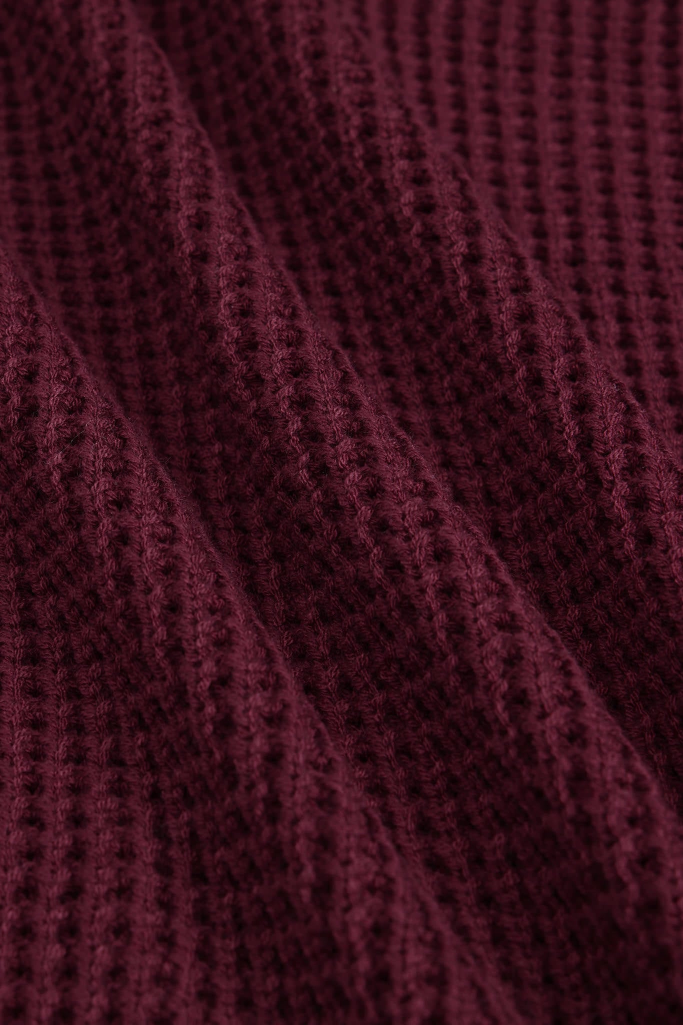 A close-up of a textured knit fabric in a deep burgundy or maroon color, showing the detailed waffle-knit pattern and soft folds.
