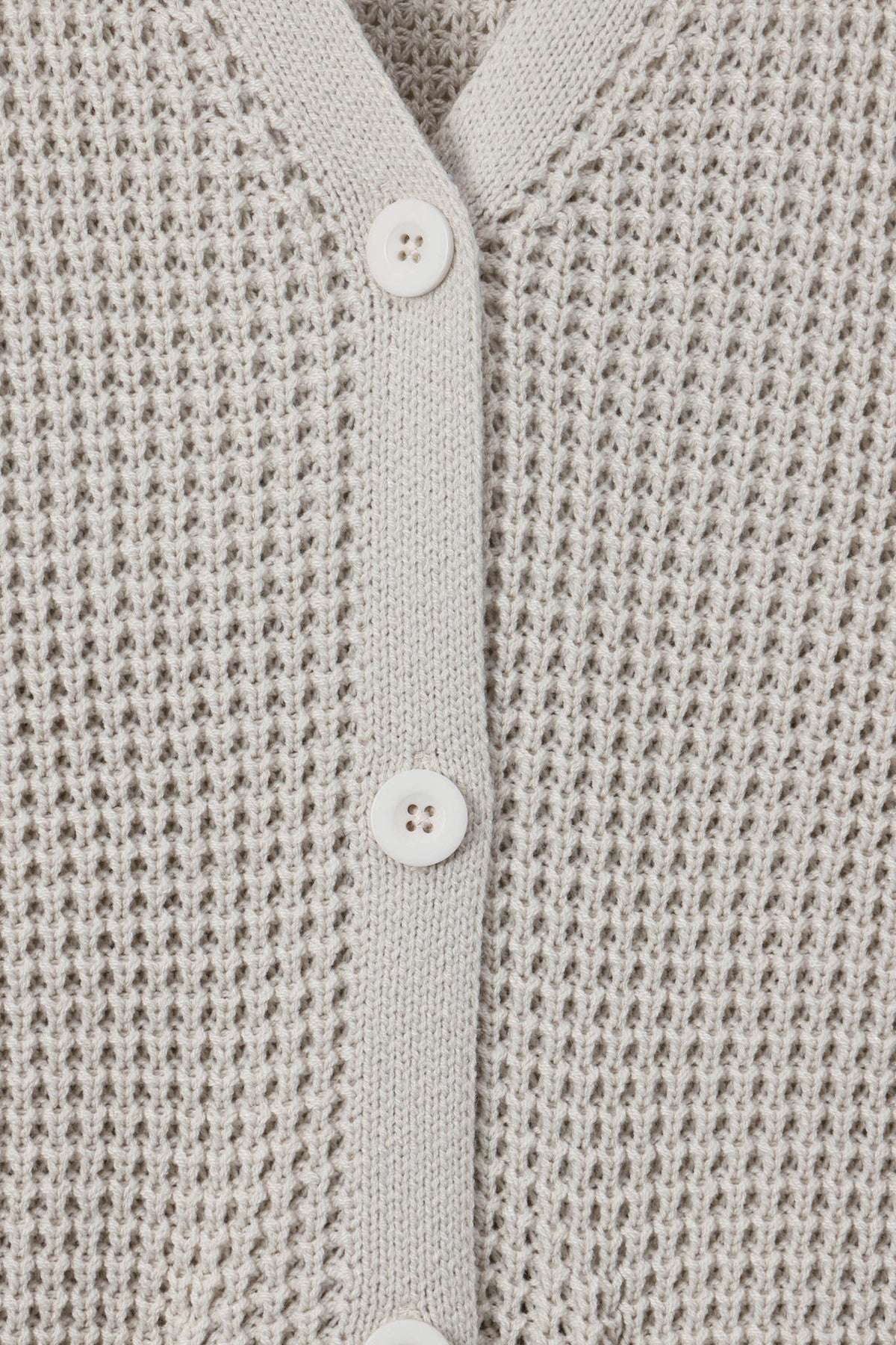 Close-up of a textured knit cardigan in a light warm gray or oatmeal tone, featuring a button placket with white buttons.