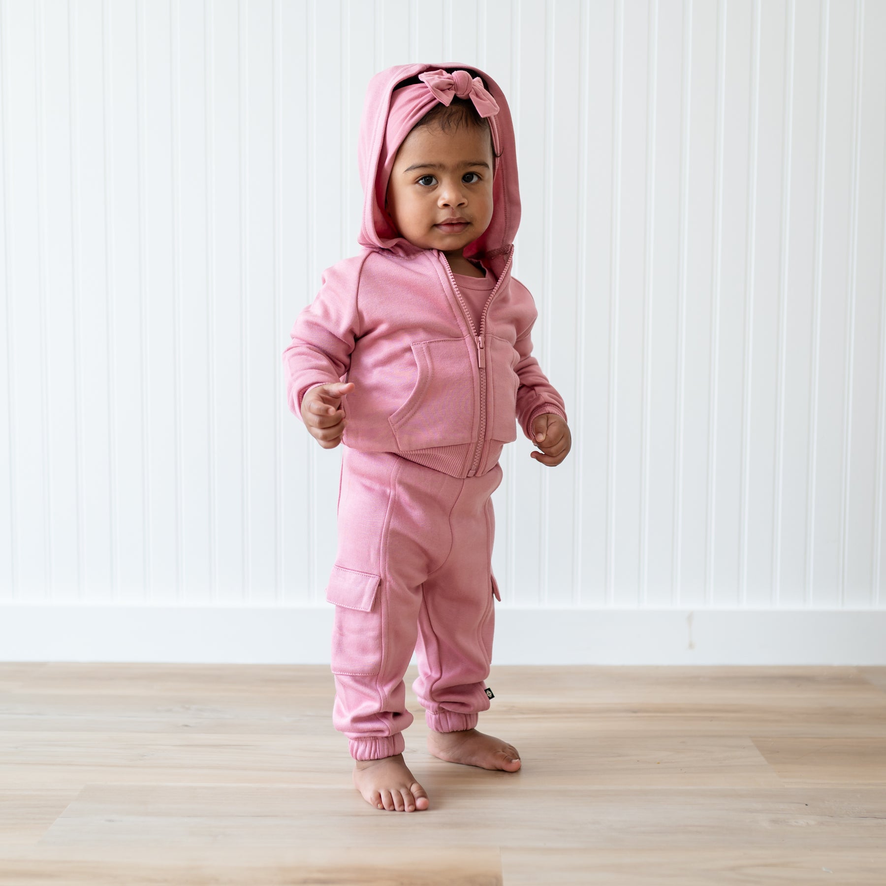 Young toddler standing wearing the Fleece Zip Up Hoodie in Apple Blossom with the hood up and matching Fleece Cargo pant standing in front of a white paneled wall
