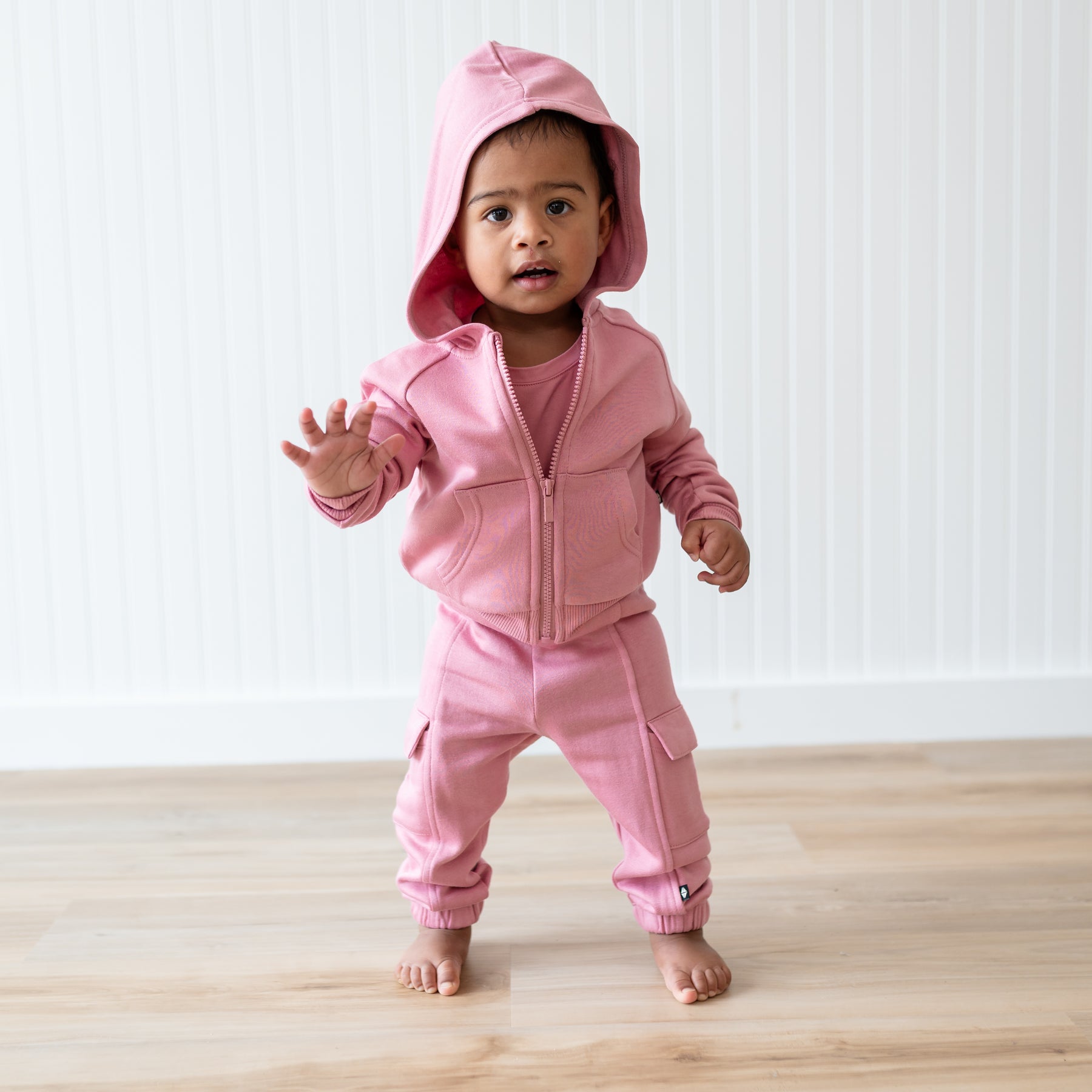 Young toddler walking wearing the Fleece Zip Up Hoodie in Apple Blossom with the hood up and the matching fleece cargo pants