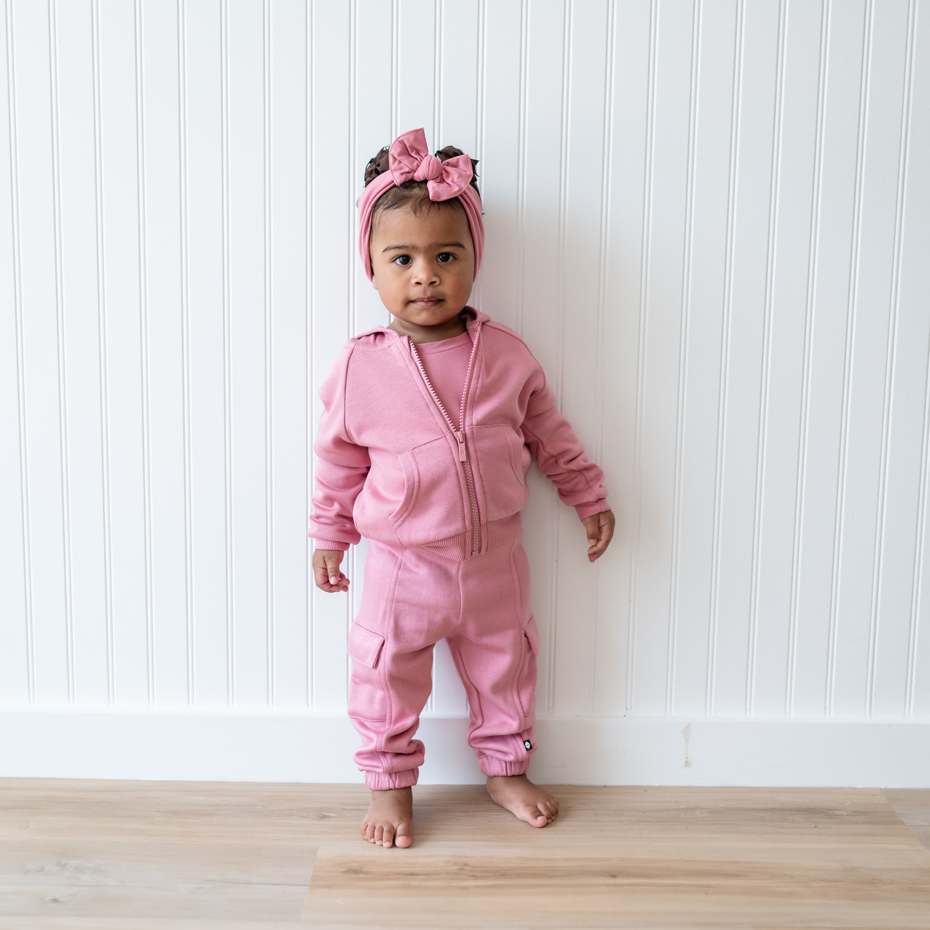 Young toddler standing in front of a white paneled wall wearing the Fleece Zip Up Hoodie in Apple Blossom and matching fleece cargo pants and Knotted bow